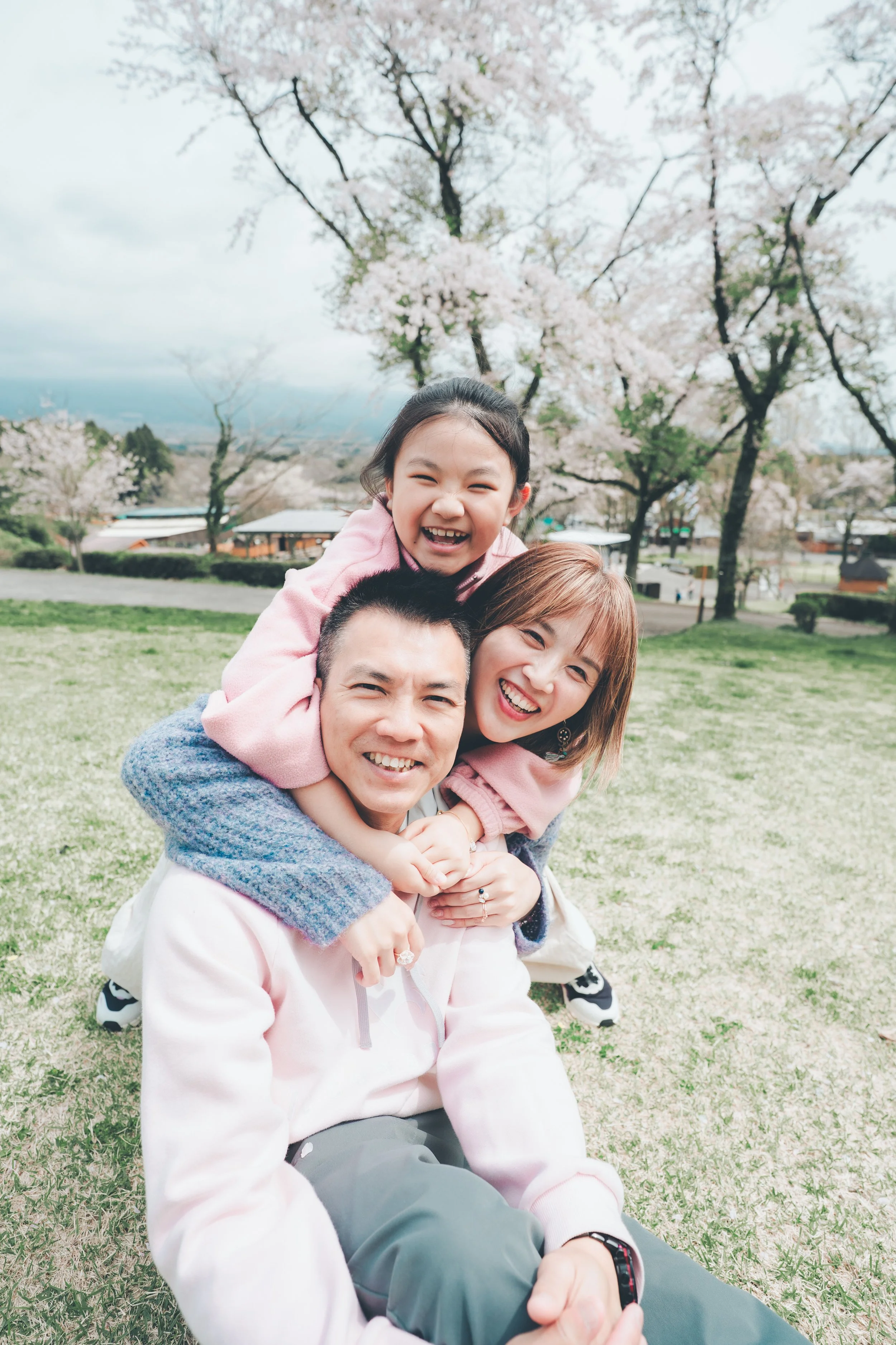 A family of three enjoying a day outdoors in a park with cherry blossom trees in bloom, with a man sitting on the grass and a woman and child on his back, all smiling happily.