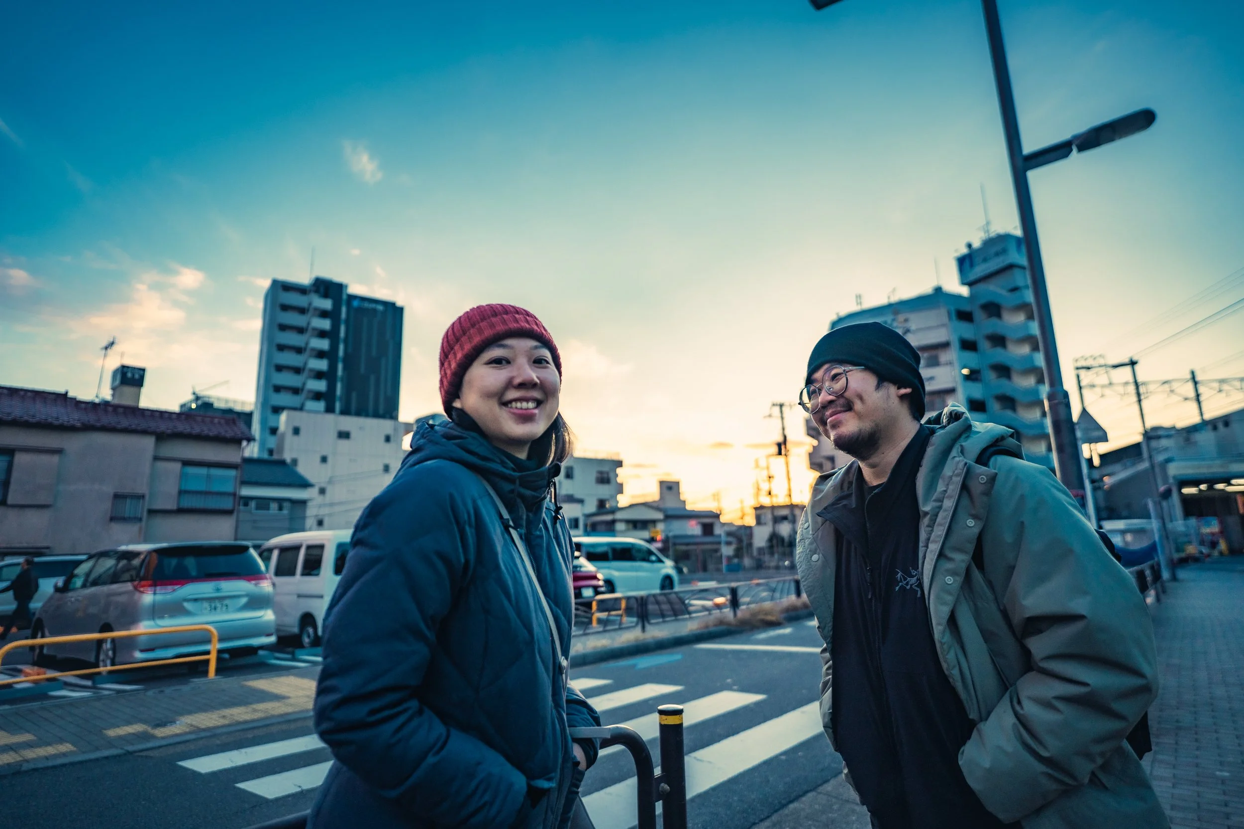 Two people standing on a city street at sunset, smiling and chatting. The woman on the left is wearing a blue jacket and red beanie, and the man on the right is wearing glasses, a black beanie, and a gray jacket.