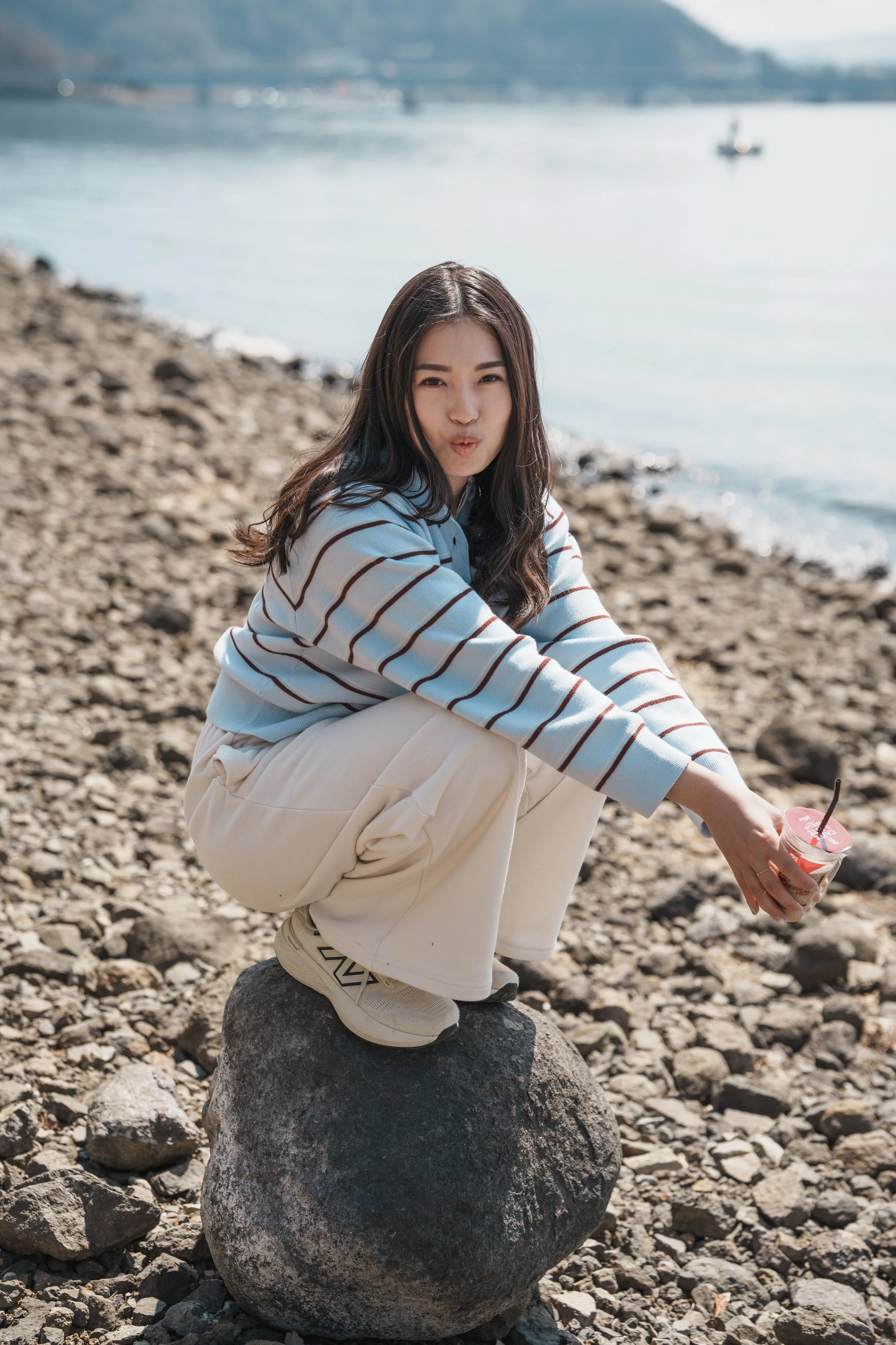 A young woman squatting on a large rock at the rocky beach, holding a cup with a straw, with water and mountains in the background.