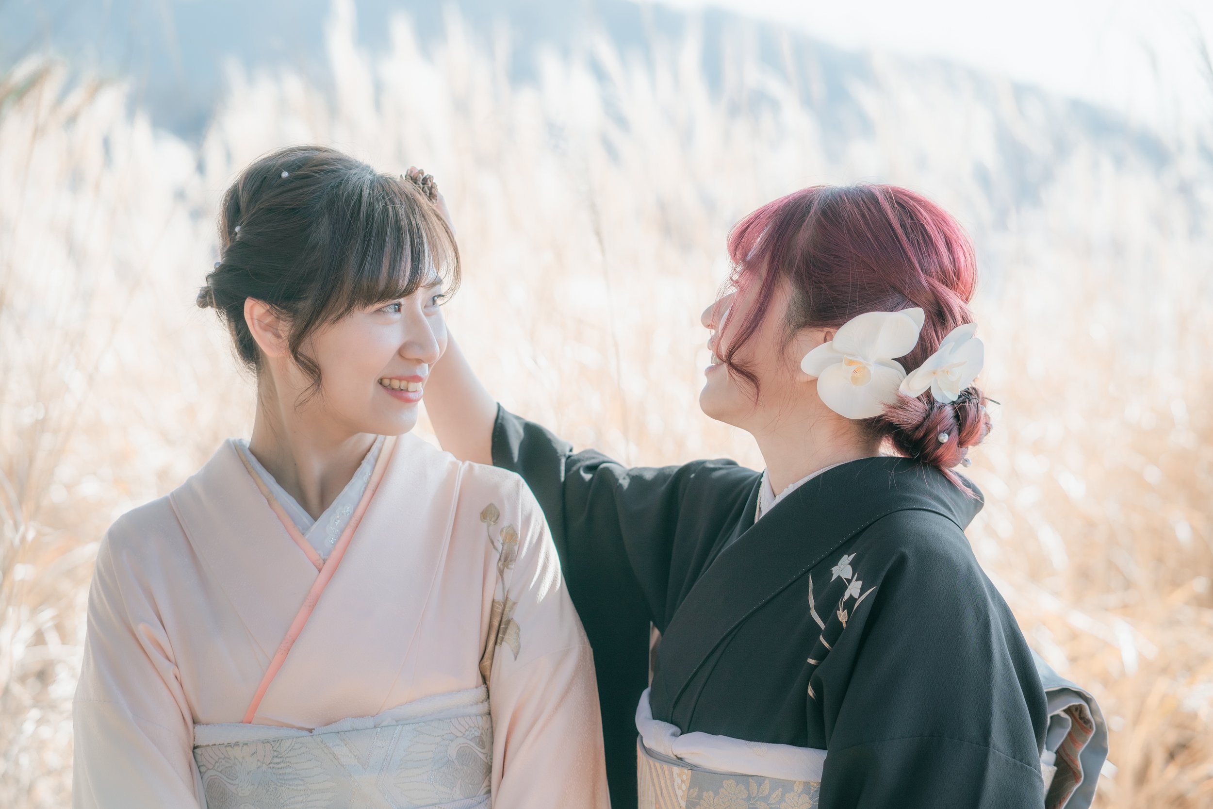 Two women dressed in traditional kimono clothing standing close together outdoors, smiling at each other with a background of tall, golden grass.