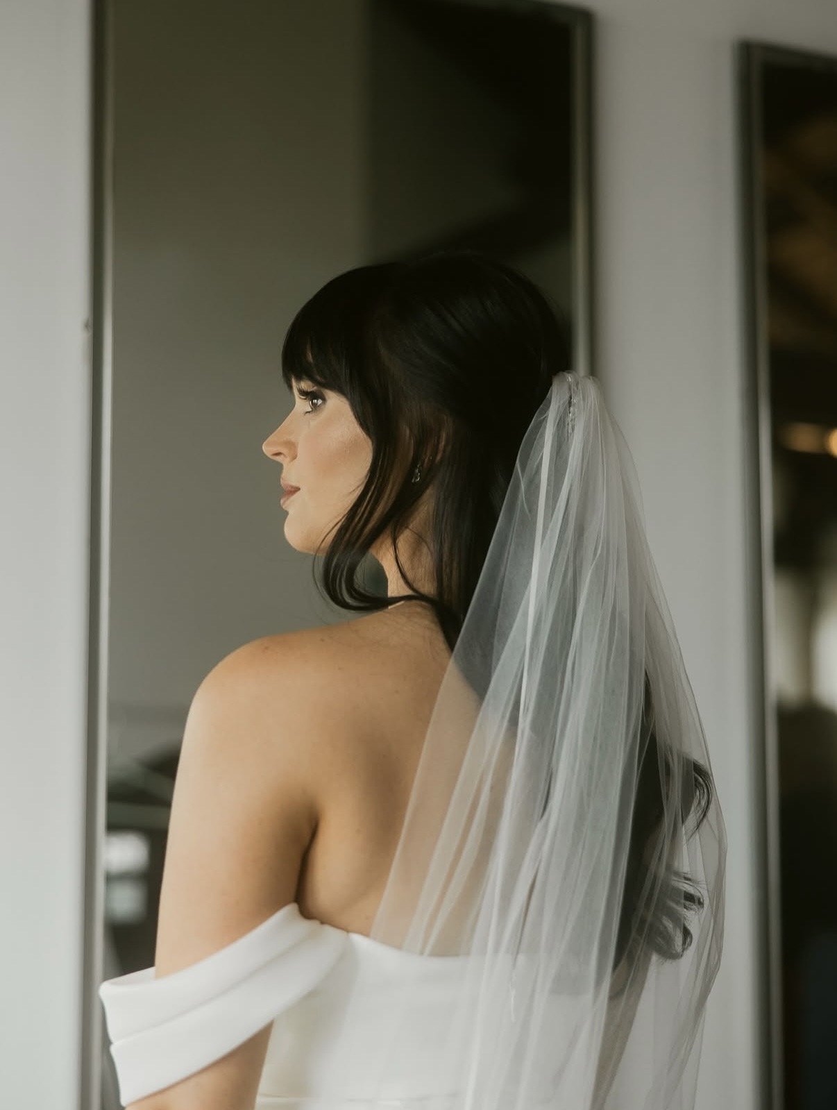 Brunette bride with warm olive skin, long soft waves and bangs wearing a sheer veil and off-the-shoulder wedding gown