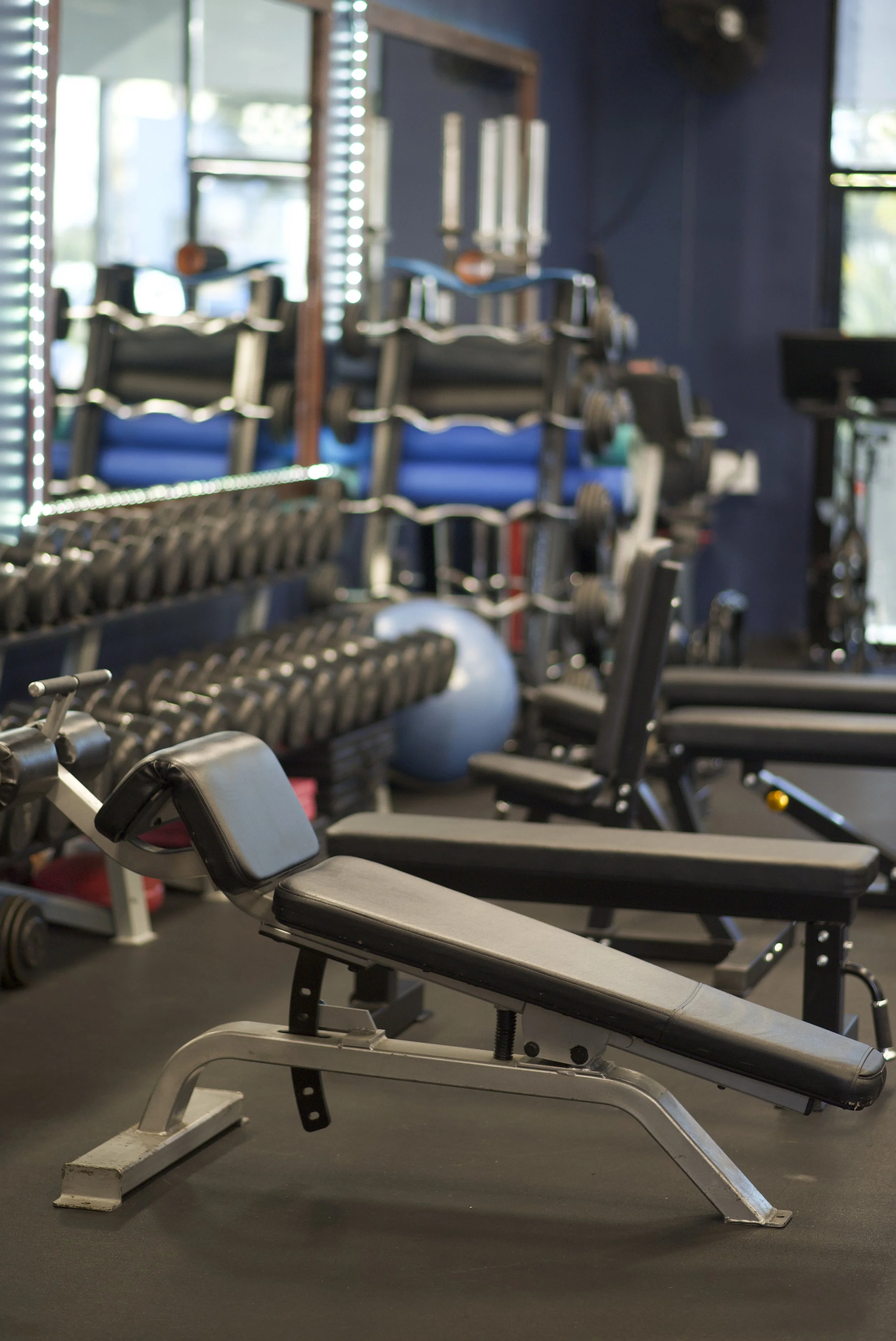 Indoor gym with workout equipment including adjustable weight benches, dumbbells, yoga mats, and a stability ball, with a mirror and window in the background.