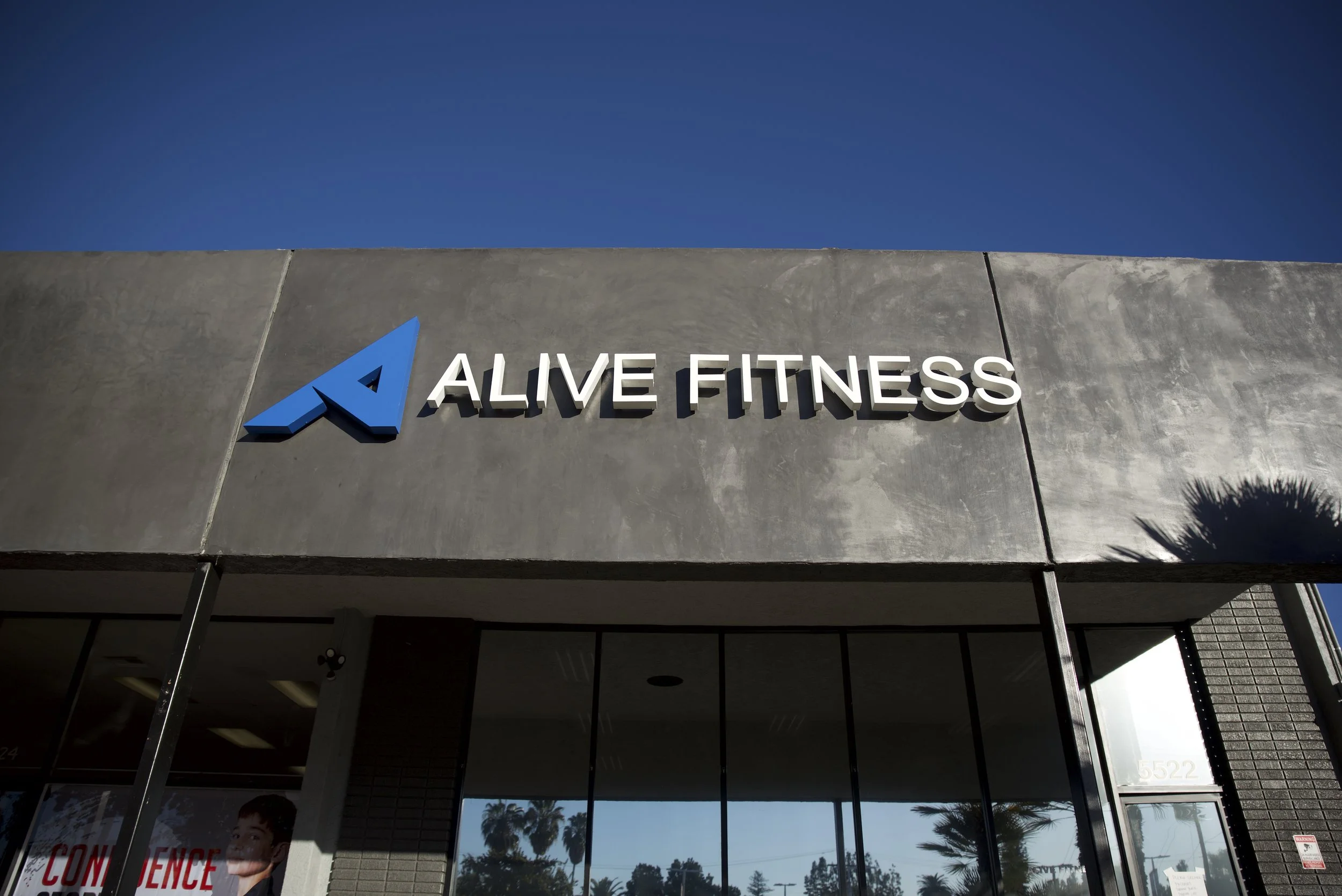 The exterior of an AlMade Fitness gym with its sign on a gray concrete wall under a blue sky, featuring a stylized blue 'A' logo and white letters spelling 'ALIVE FITNESS'.
