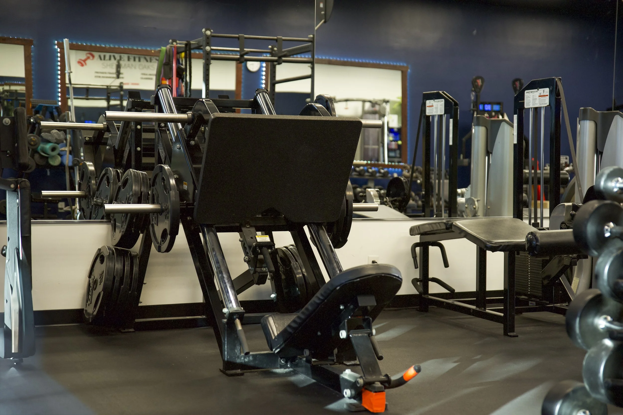 Fitness gym interior with various exercise equipment including a leg press machine, a bench, and dumbbells, with mirrors and dim lighting.