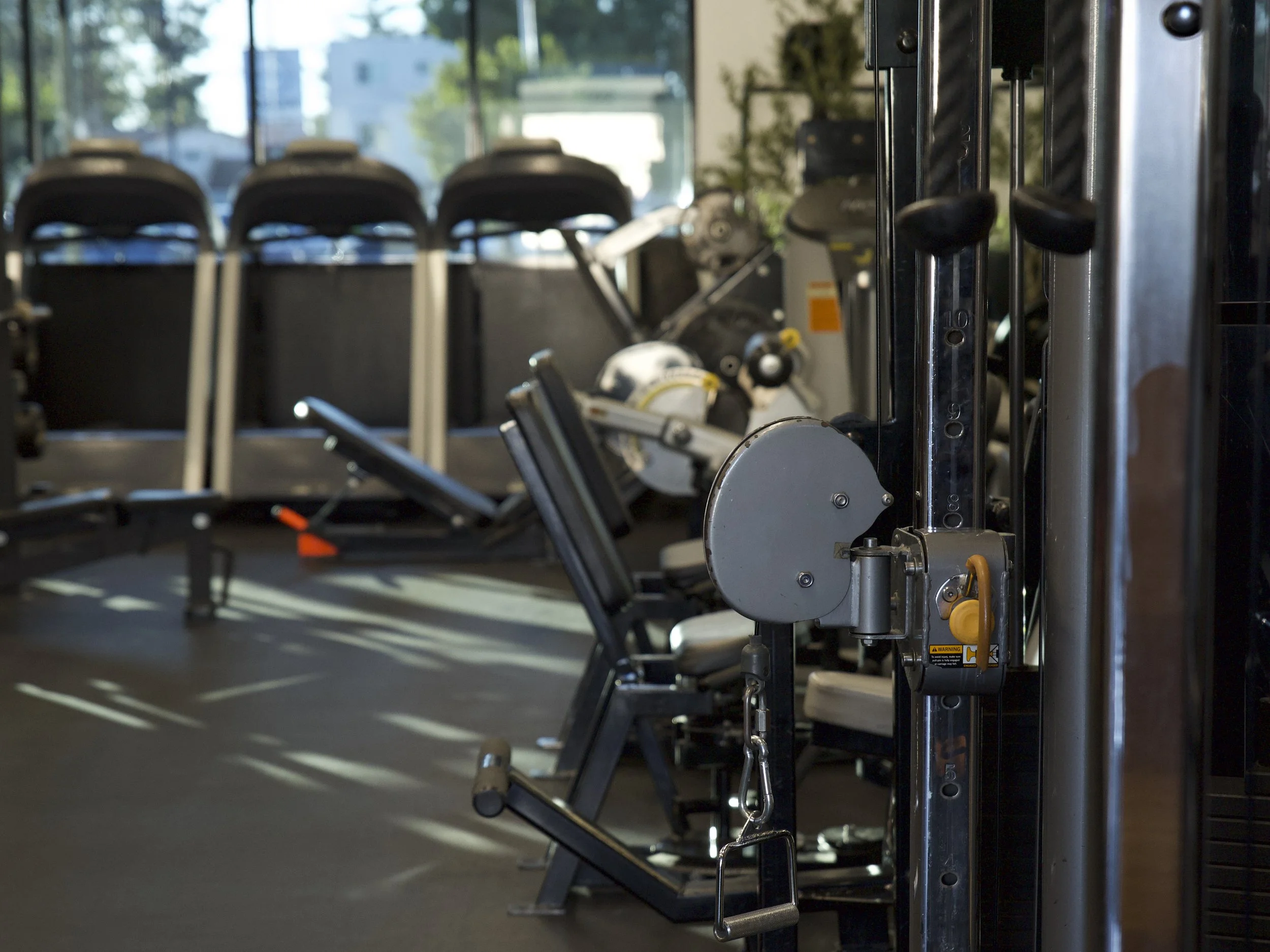 Close-up of gym equipment, including a cable machine in the foreground, with treadmills and rowing machines in the background inside a fitness center with large windows showing trees and buildings outside.
