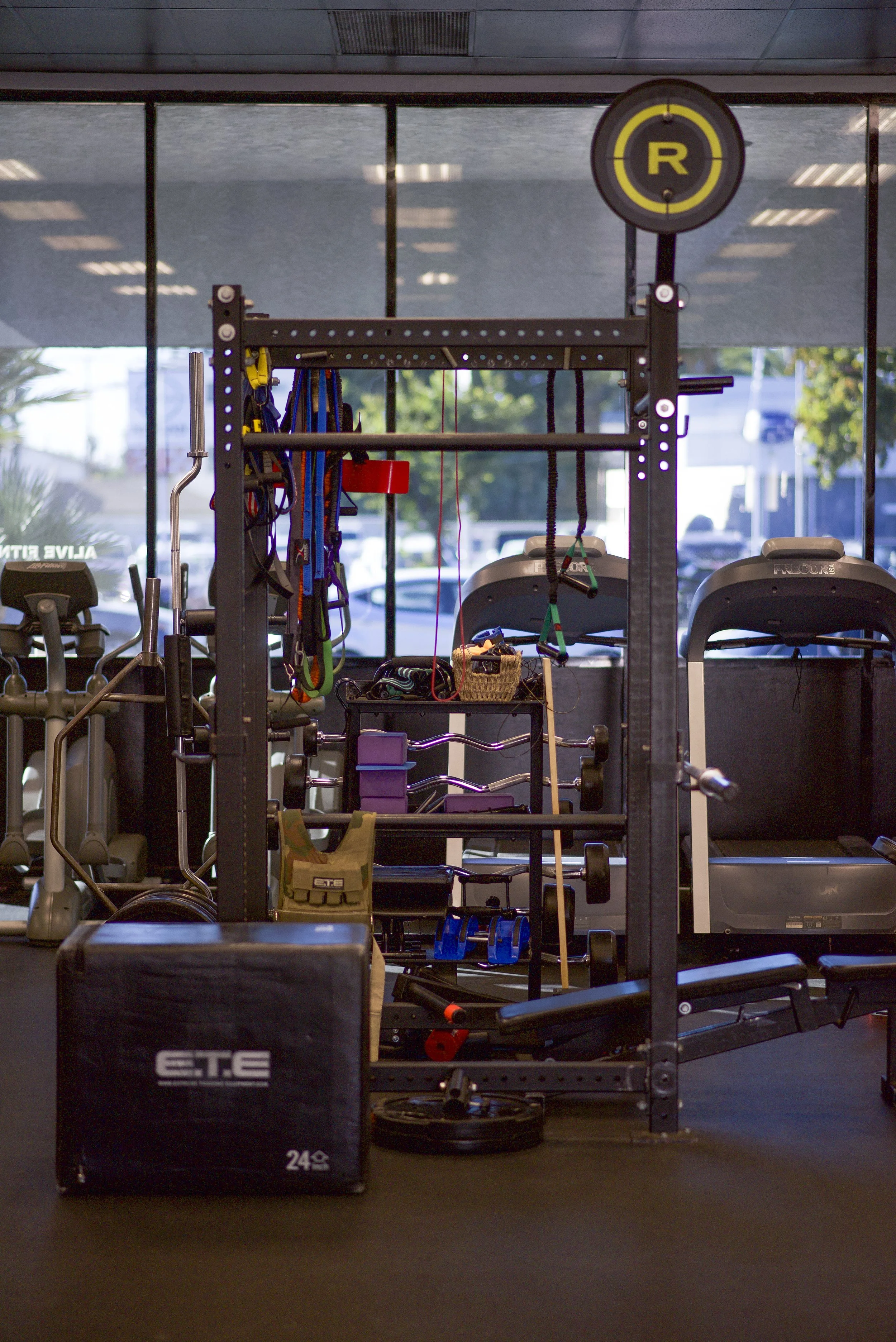Empty gym with workout equipment, including a squat rack, dumbbells, a plyometric box, treadmills, and various resistance bands, with large windows in the background.