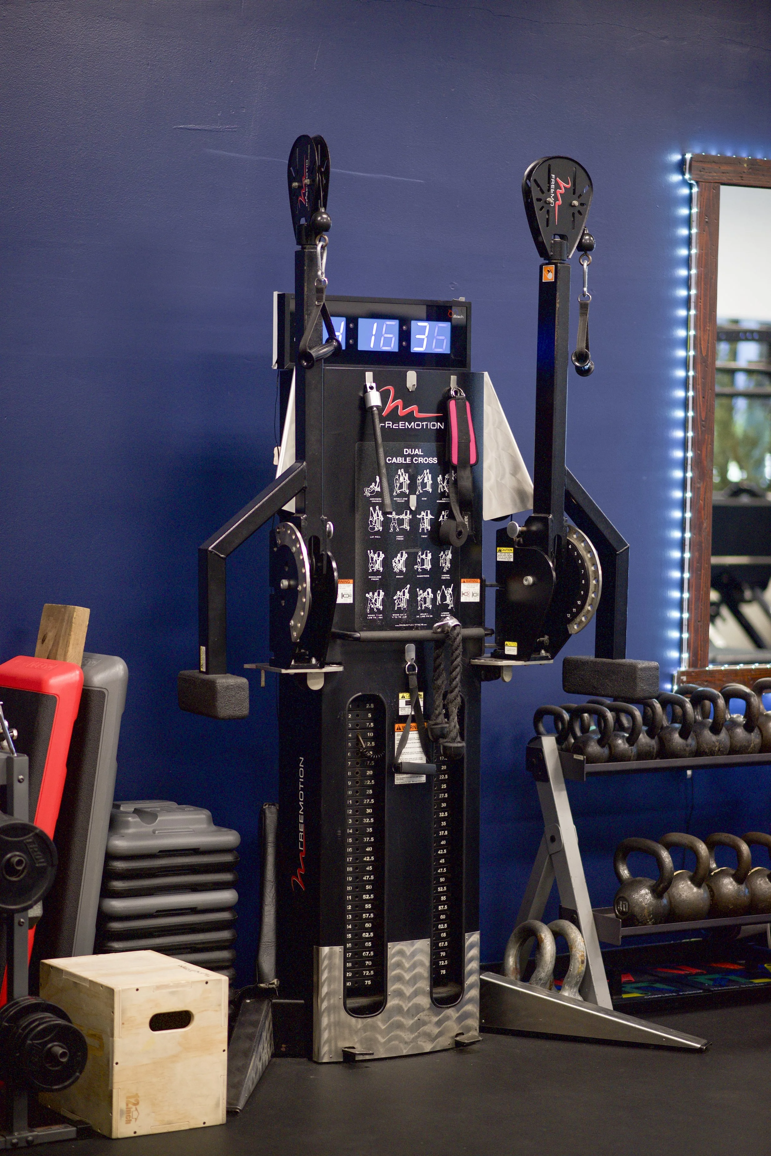 A gym with various workout equipment, including a dual cable cross machine with a digital clock showing 16:36, and kettlebells on a rack.