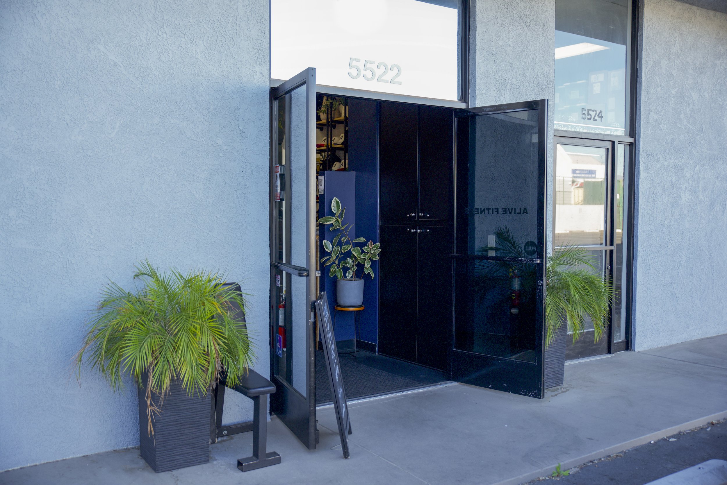 Office building entrance with open black glass doors, potted plants on either side, and street view through glass windows.