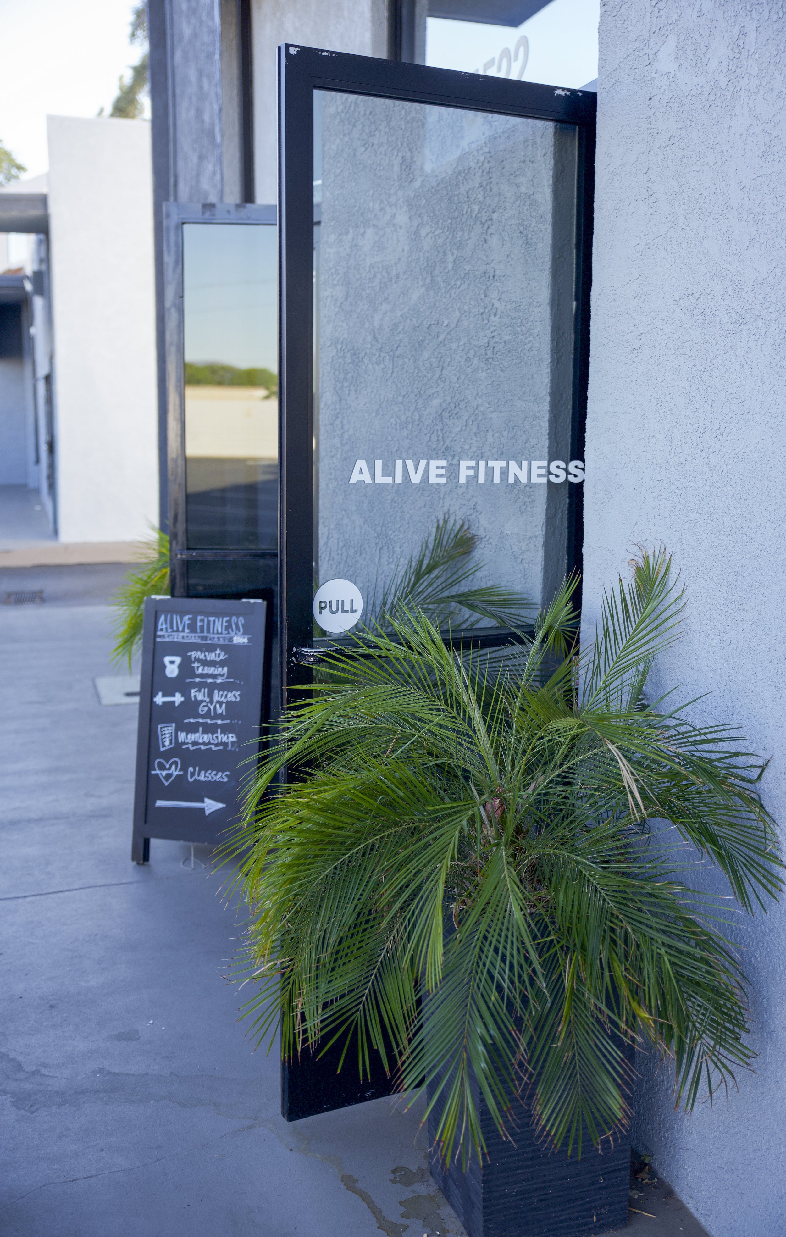 The entrance to ALIVE FITNESS gym with a glass door, a potted palm plant, and a chalkboard sign listing gym features.