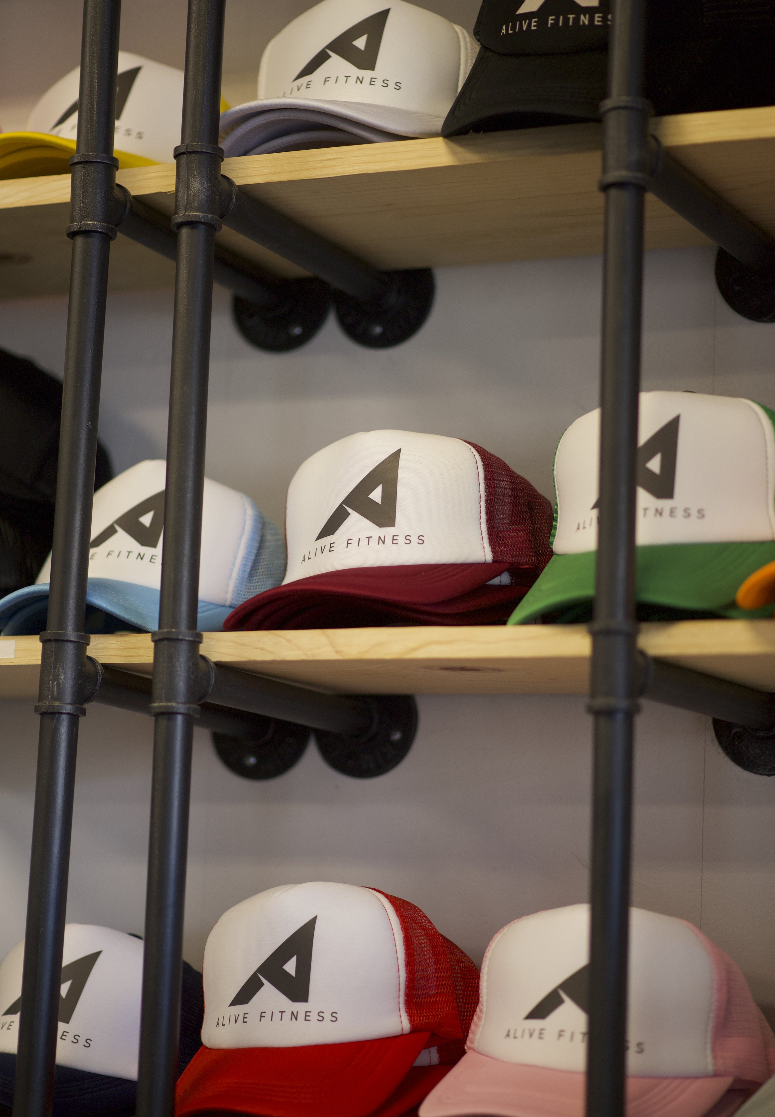 A display of colorful trucker hats with the brand name 'Alive Fitness' and a stylized 'A' logo on the front, arranged on wooden shelves behind a black metal frame.