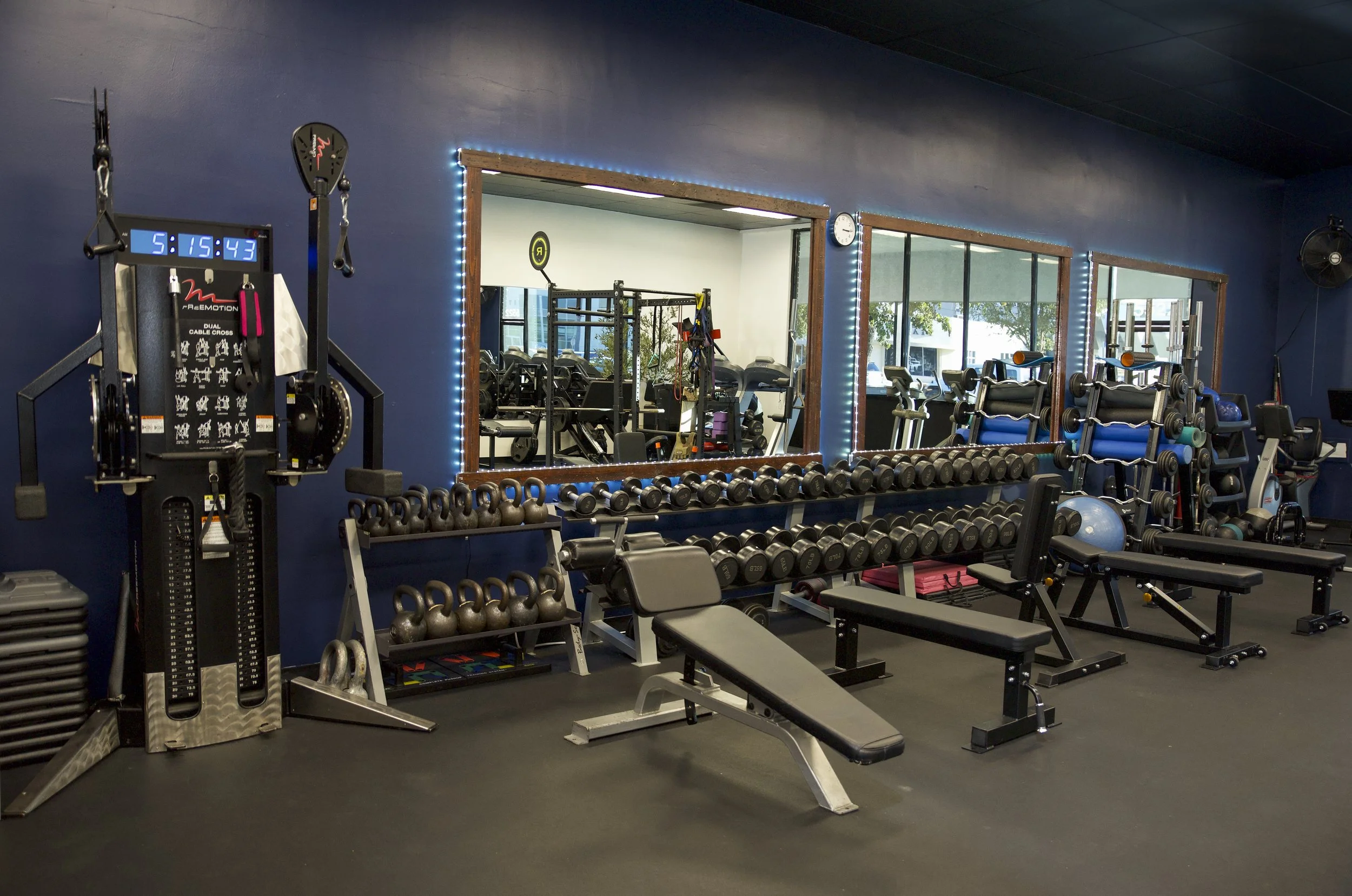 Indoor gym with weight racks, benches, kettlebells, and mirrors, featuring blue walls and large windows.