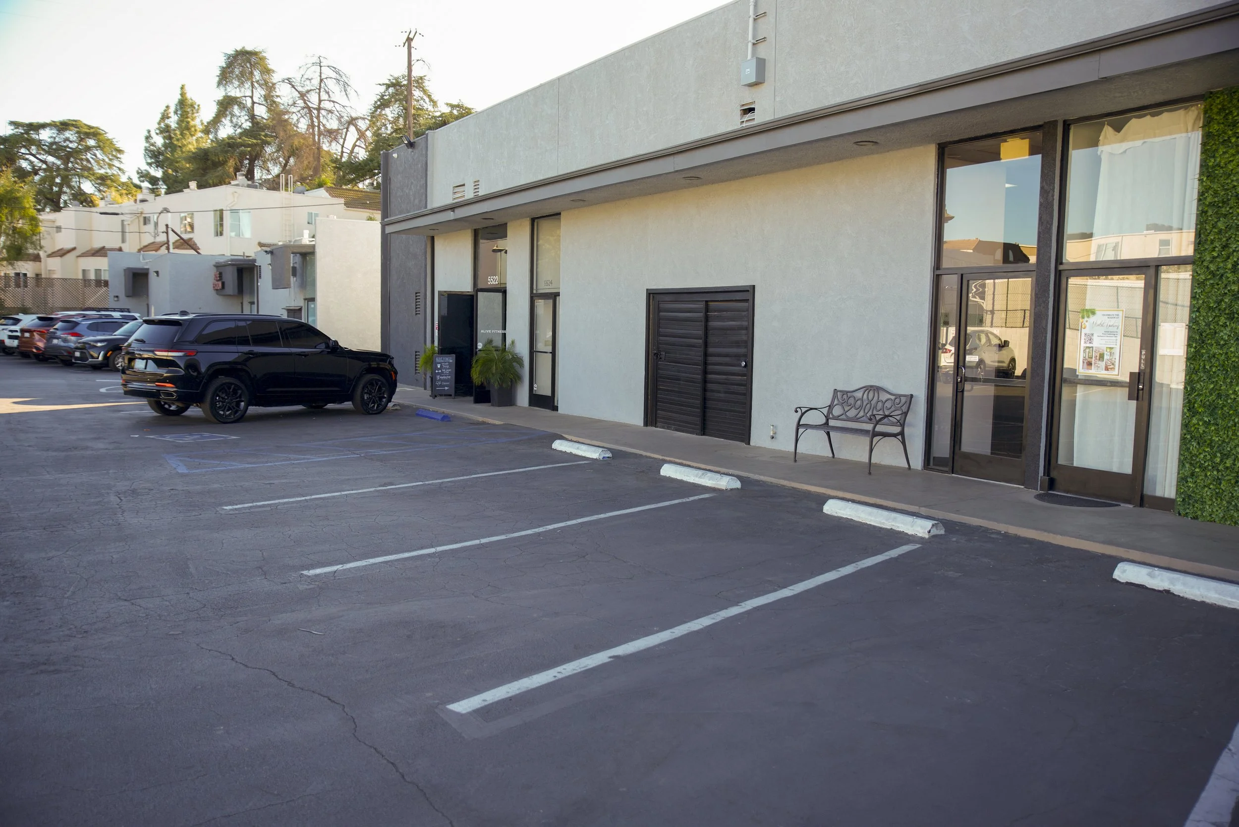 Empty parking lot in front of a commercial building with parked cars and a bench by the entrance.