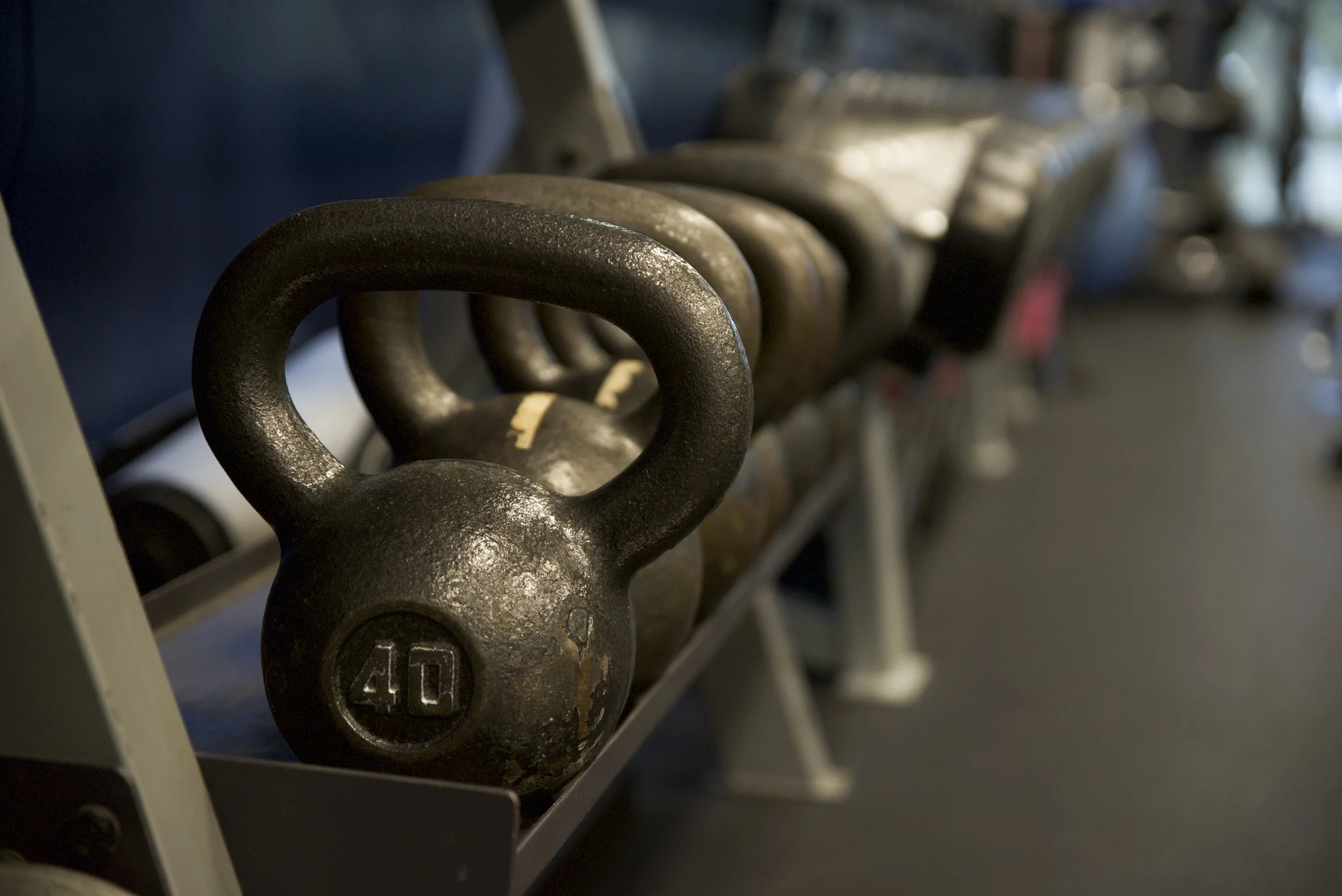 Row of black cast iron kettlebells with varying weights on a metal rack in a gym.