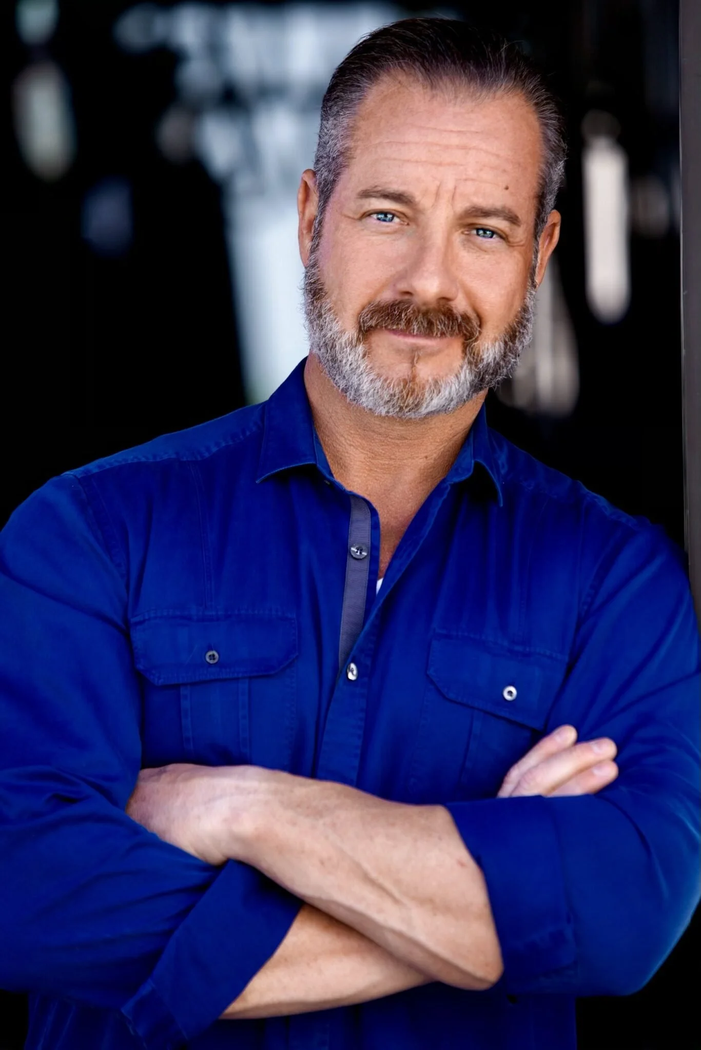 A middle-aged man with gray hair and beard, wearing a blue button-up shirt, standing outdoors with arms crossed and smiling at the camera.