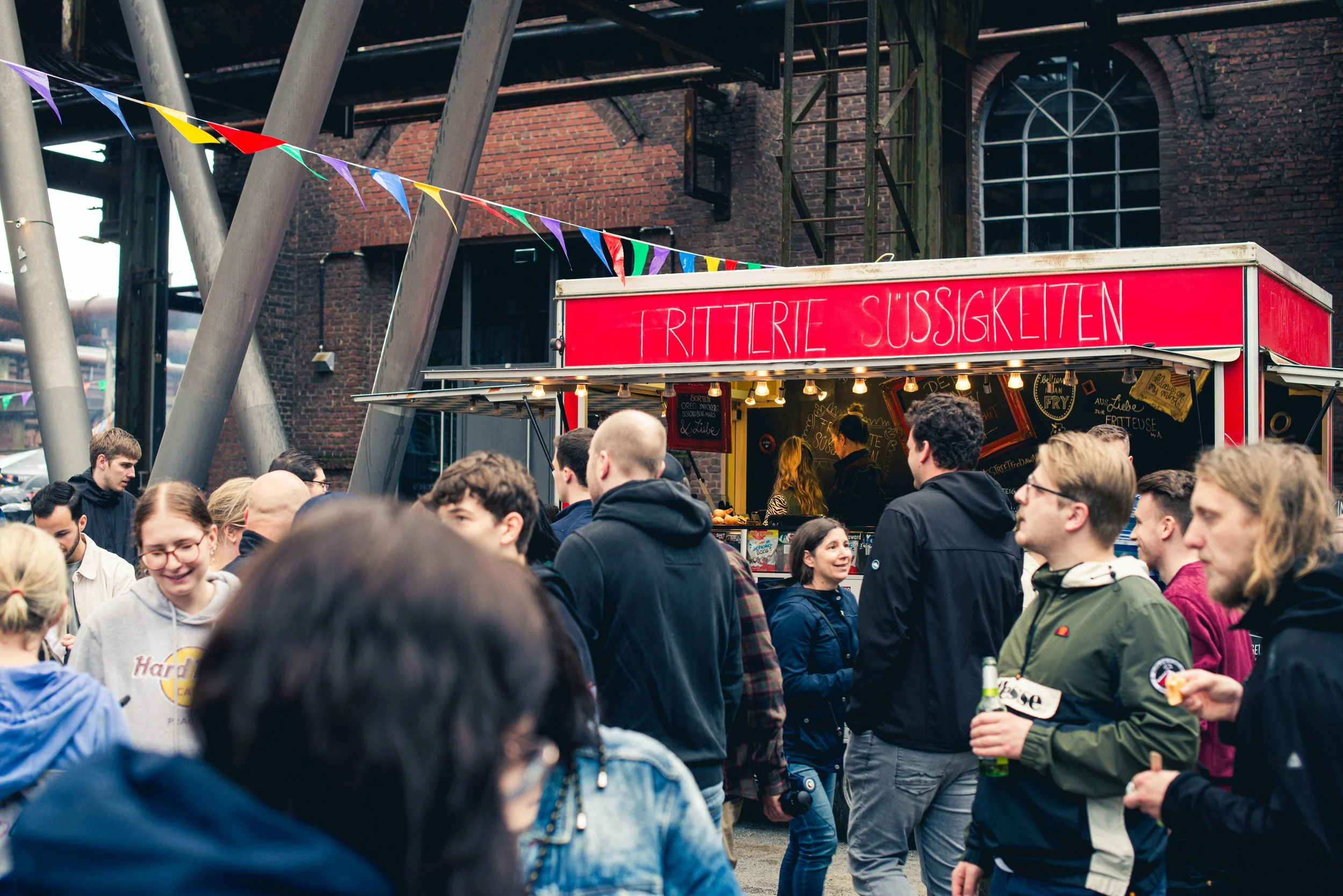 Menschenmenge bei einem Straßenfest mit bunten Fähnchen und einem Verkaufsstand gegen eine Backsteinwand.