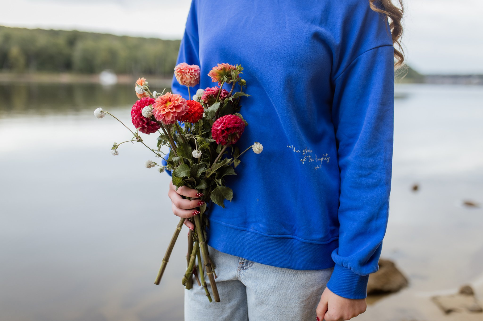 Person wearing a blue sweatshirt holding a bouquet of pink and white flowers by a lake.