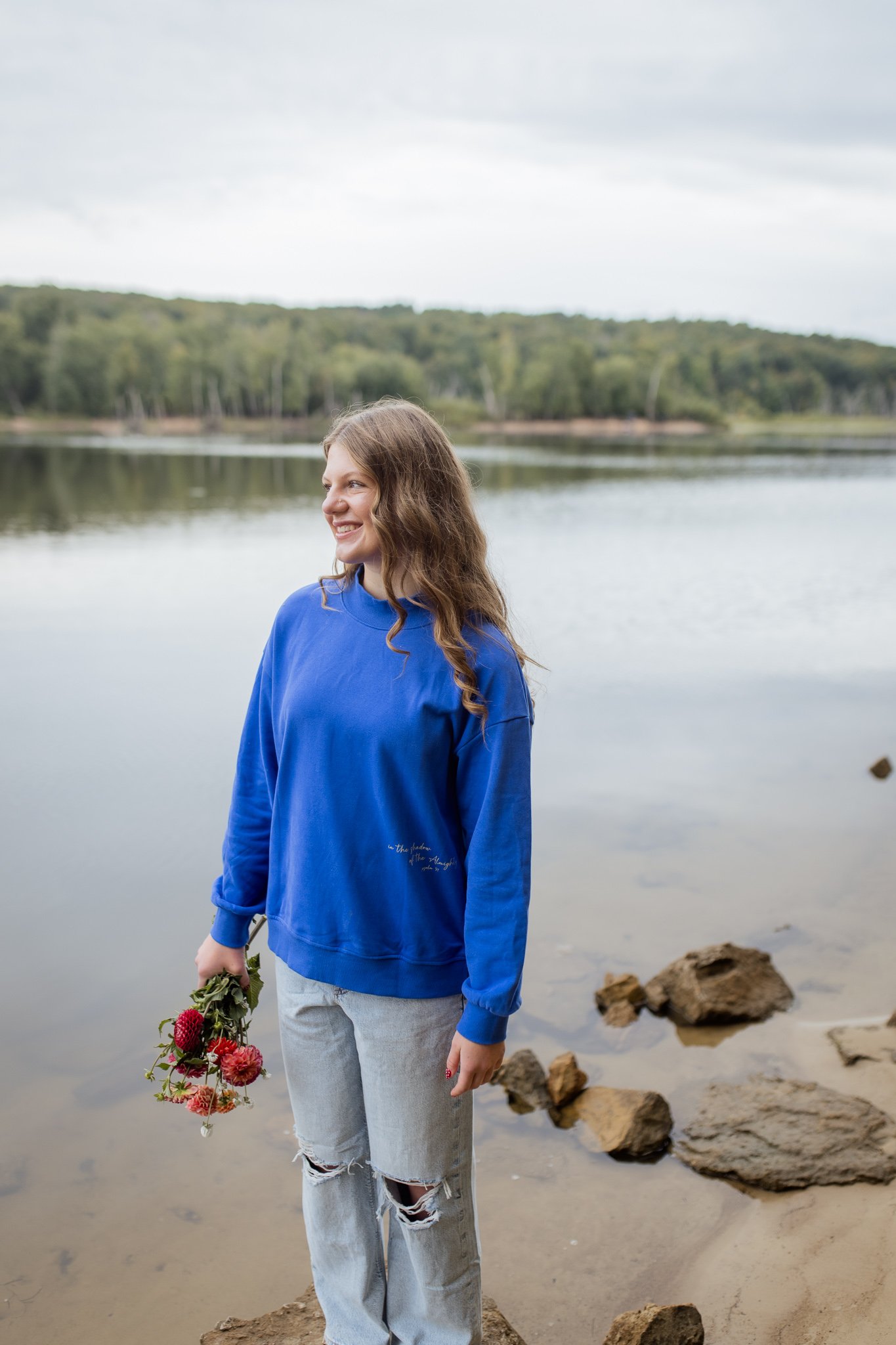 A woman with long wavy hair smiling and holding a bouquet of flowers near a body of water with rocks and a forested shoreline in the background during a fall line christian photoshoot featuring the mockneck sweater designed with Shadow of the Almight