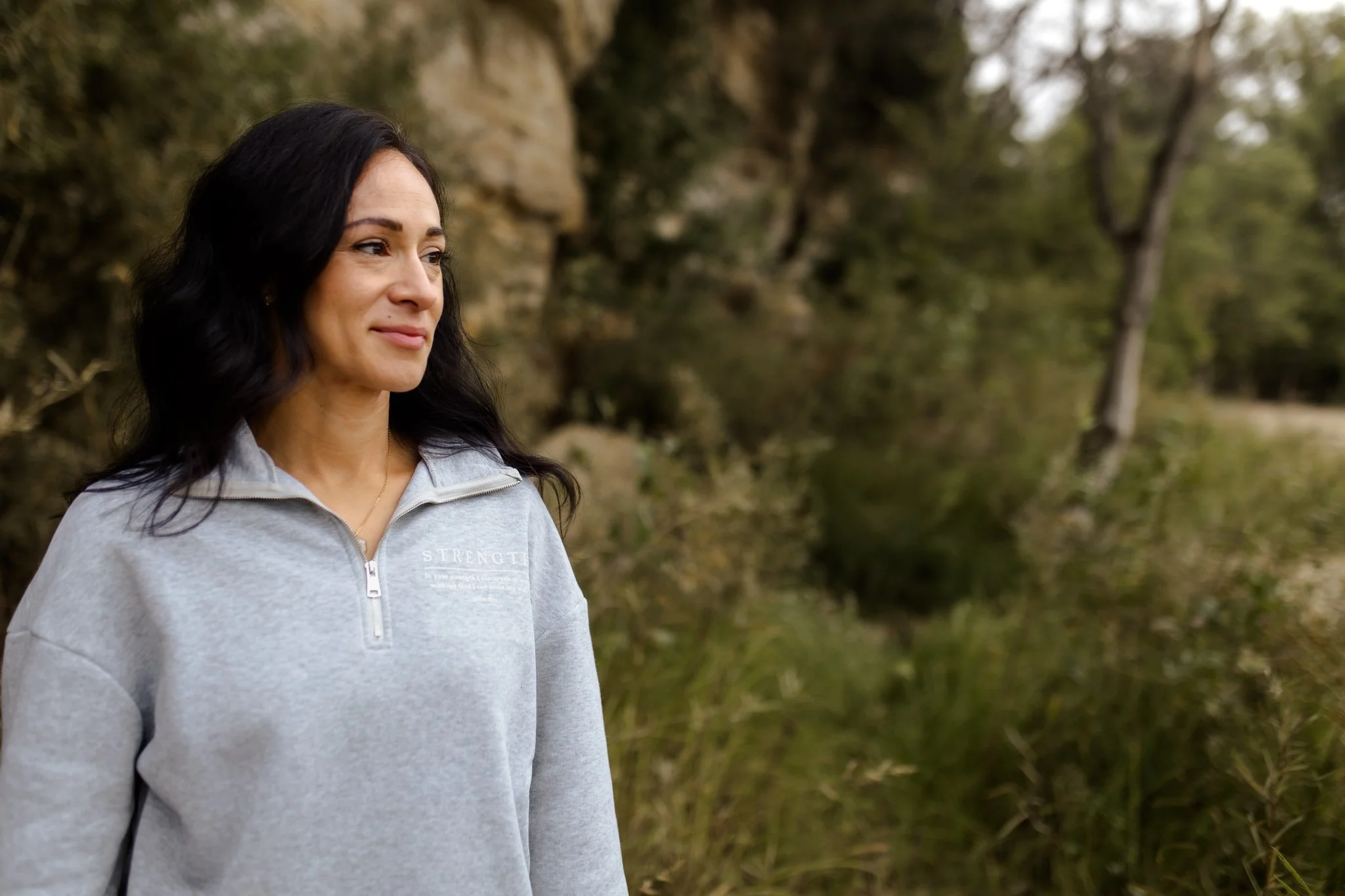 A woman with long dark hair wearing a gray zip-up sweatshirt with the words STRENGTH from psalm 18:29 outdoors with trees and a body of water in the background.