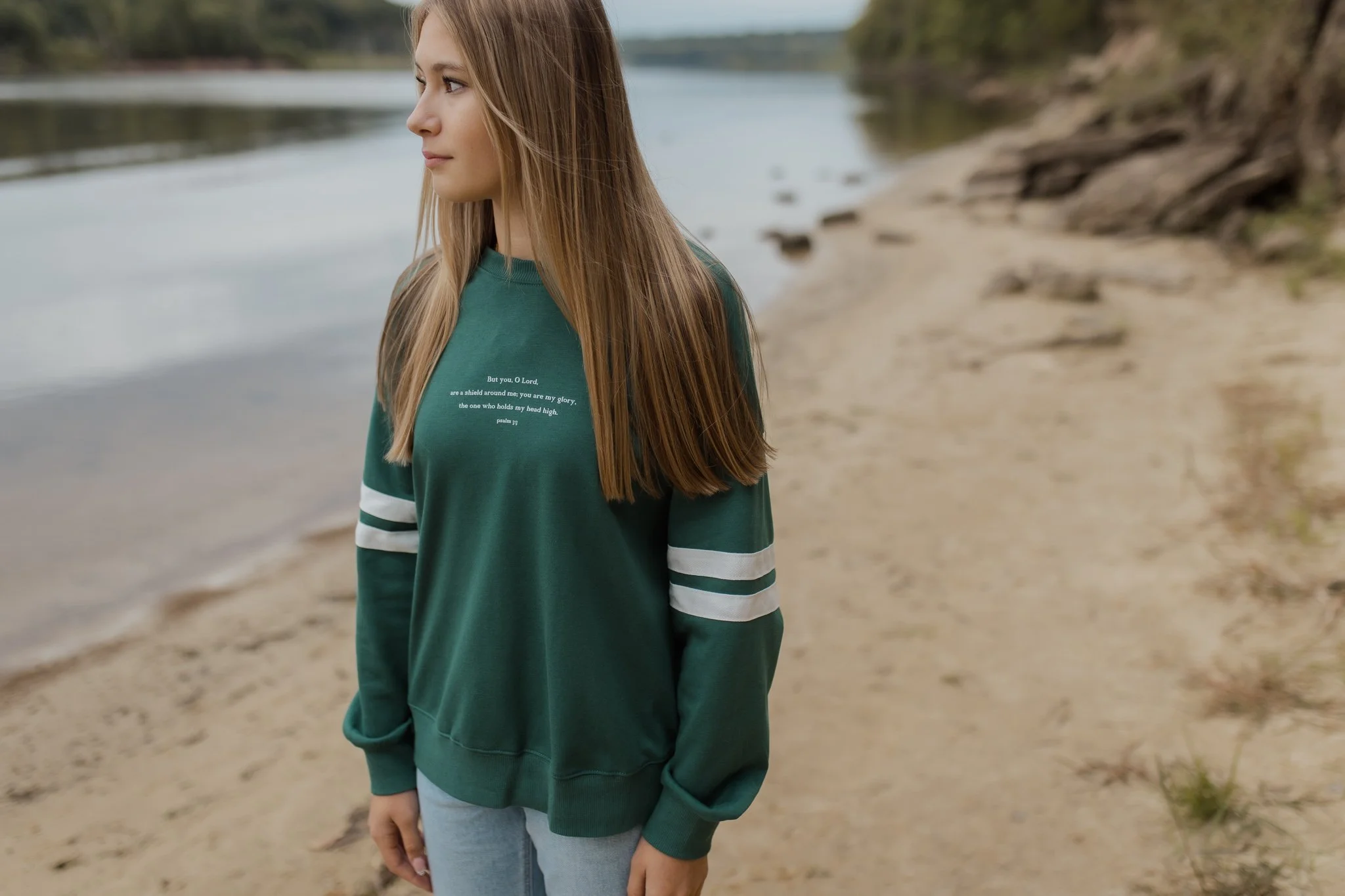 A young woman with long blonde hair standing near a lake on a sandy beach, wearing a green sweatshirt with white text and gray pants. The background features calm water and a distant shoreline during a fall line christian photoshoot
