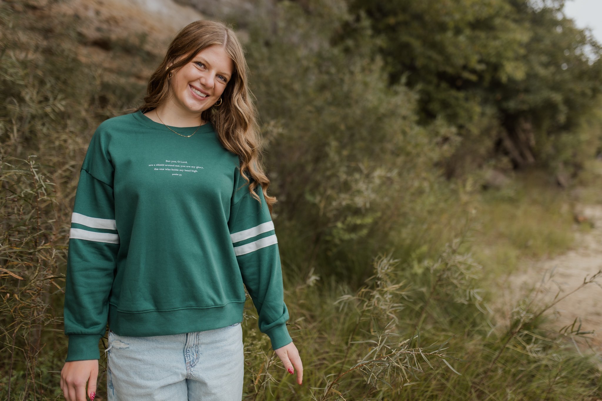 A young woman with long wavy hair smiling outdoors, wearing a green sweatshirt with white stripes on the sleeves and text on the front from psalm 3:3, and light blue jeans, standing on a nature trail surrounded by green bushes and trees.
