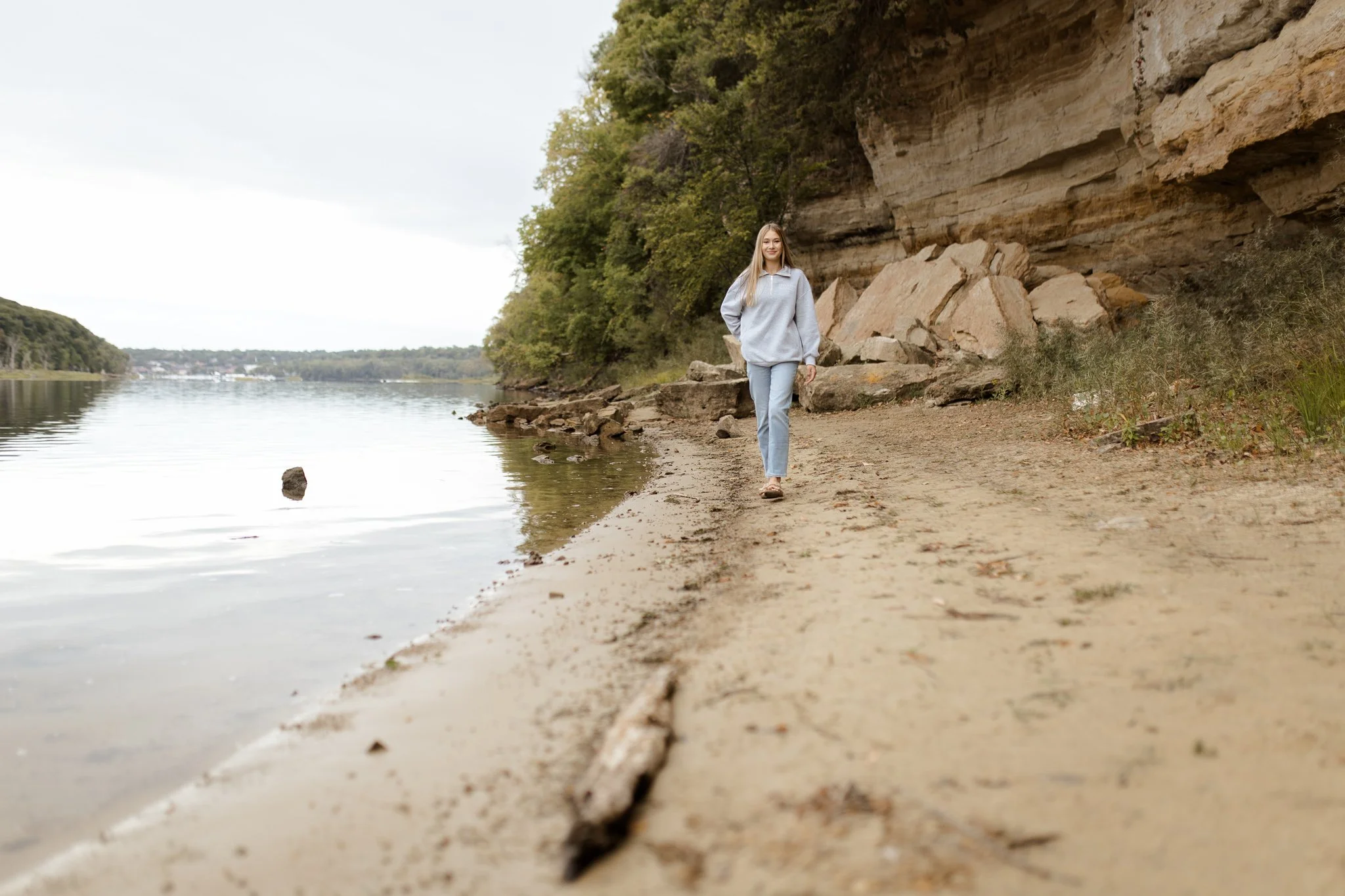 A young woman with long blonde hair walking along a sandy beach near a rocky cliff, with a body of water on her left and trees in the background.