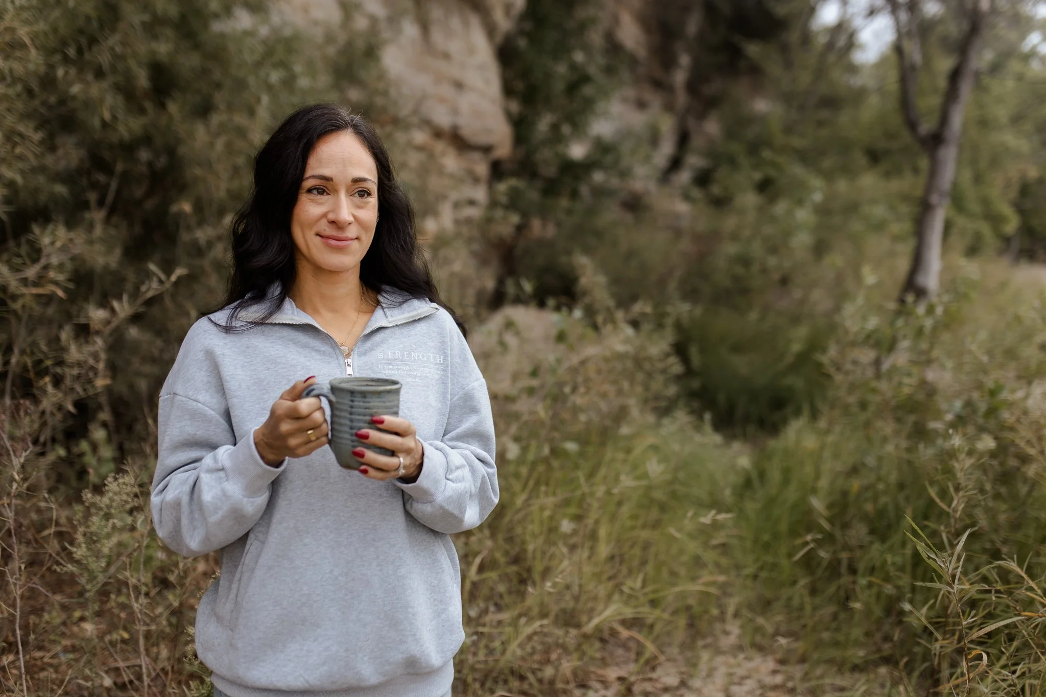 A woman with long dark hair holding a mug outdoors in a natural setting with trees and foliage for the fall line photoshoot of a christian clothing brand showcasing strength and the bible verse psalm 18:29
