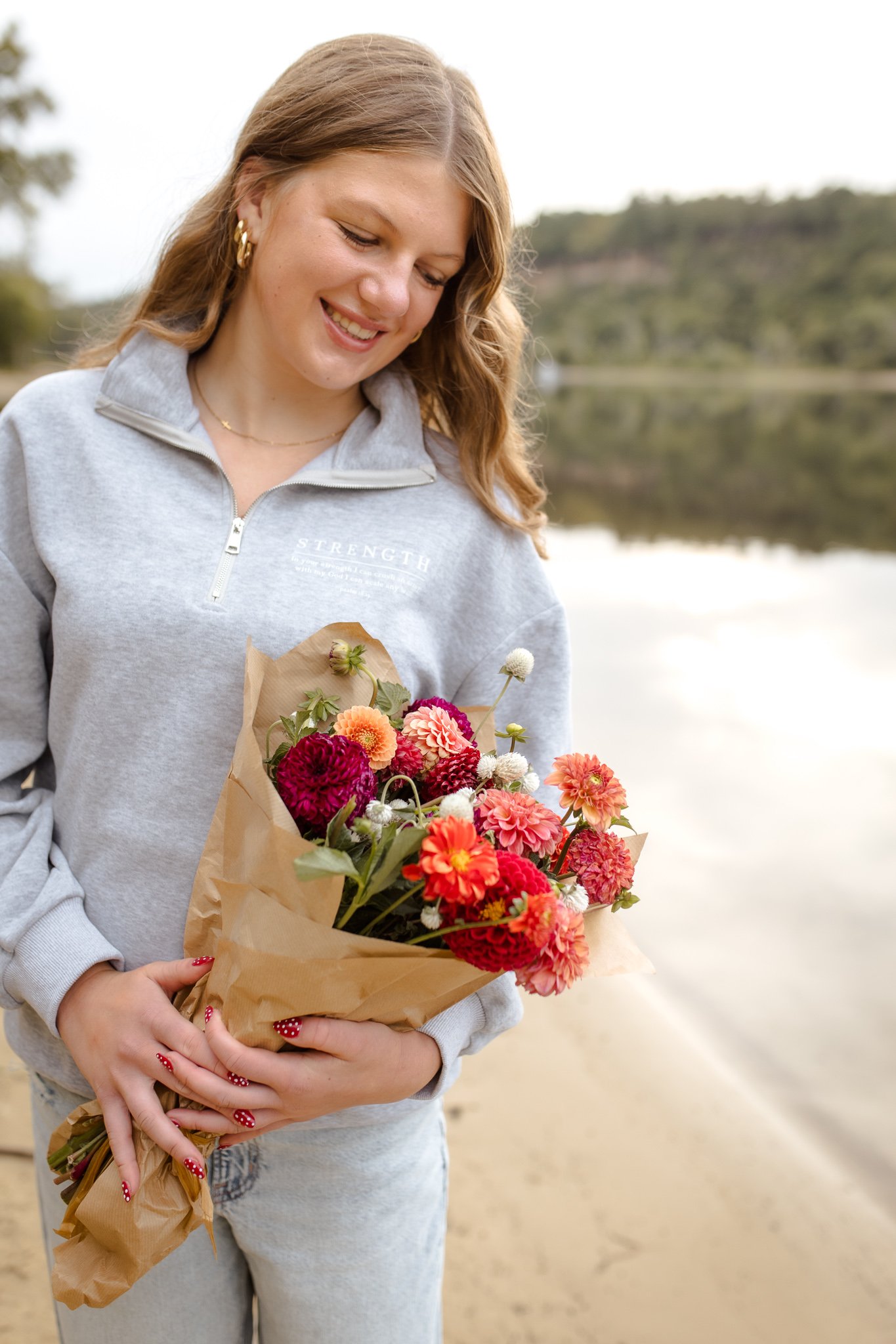A young woman with strawberry blonde hair, wearing a gray zip-up sweatshirt with the bible verse psalm 18:29, holding a bouquet of colorful flowers wrapped in brown paper, standing outdoors near a body of water with trees and hills in the background.