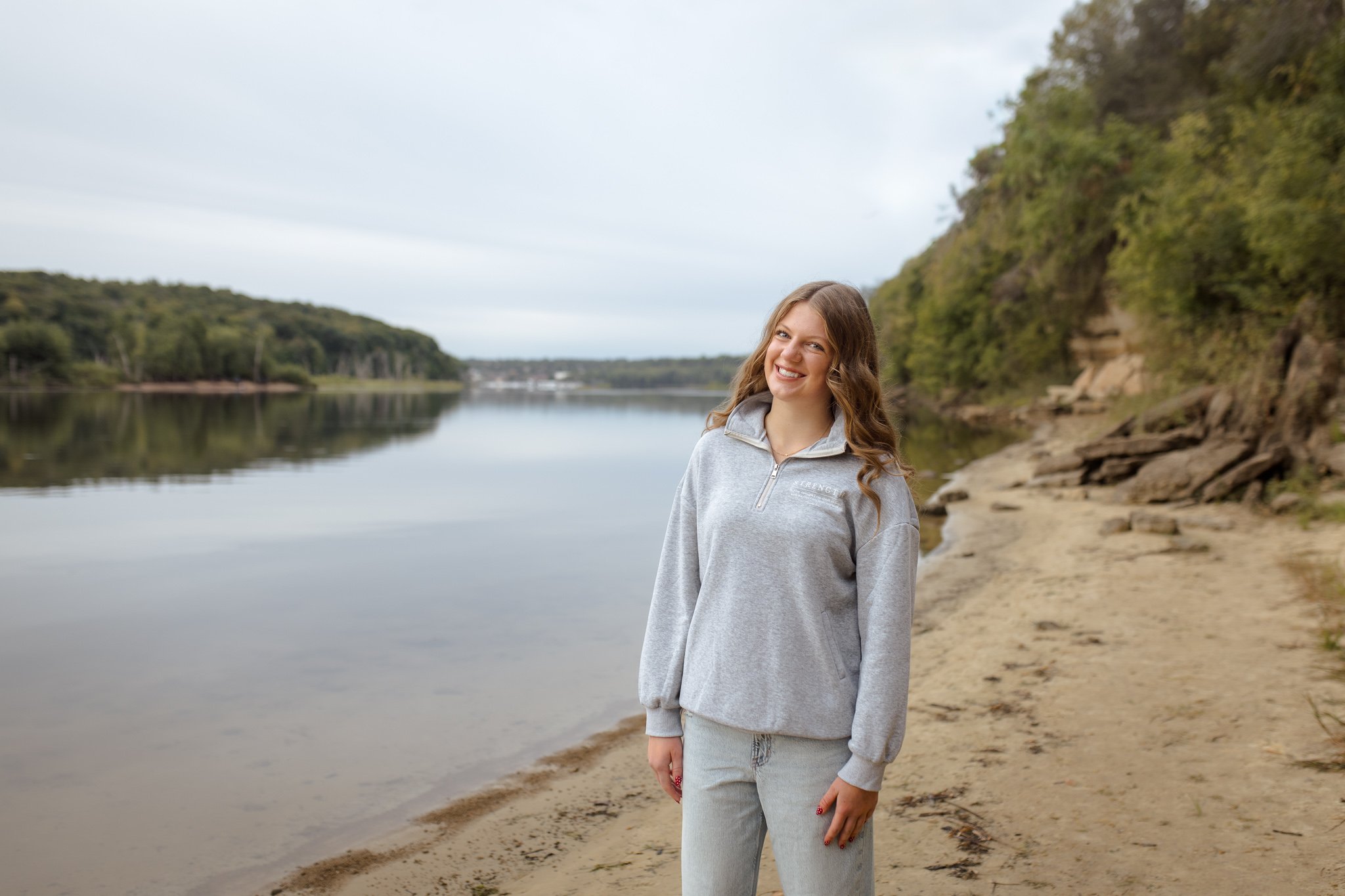 A young woman with wavy brown hair smiling while standing on a sandy beach near a calm river, with green hills and trees in the background under a cloudy sky.