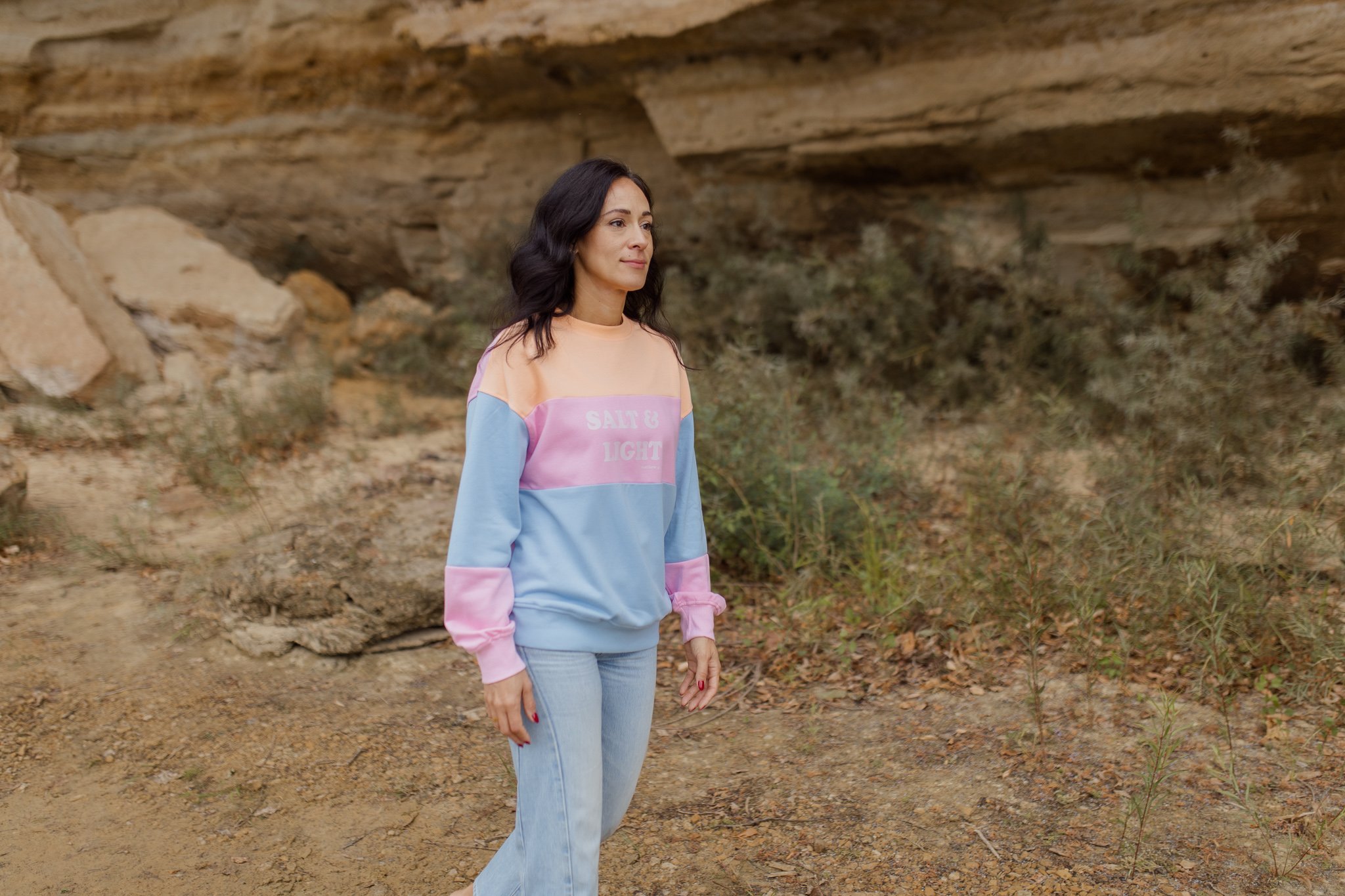 A woman with black hair and fair skin walking outdoors in a natural setting with rocks and bushes and a large rock formation overhead during a fall line photoshoot for a christian clothing company featuring a colorblock sweater with salt and light