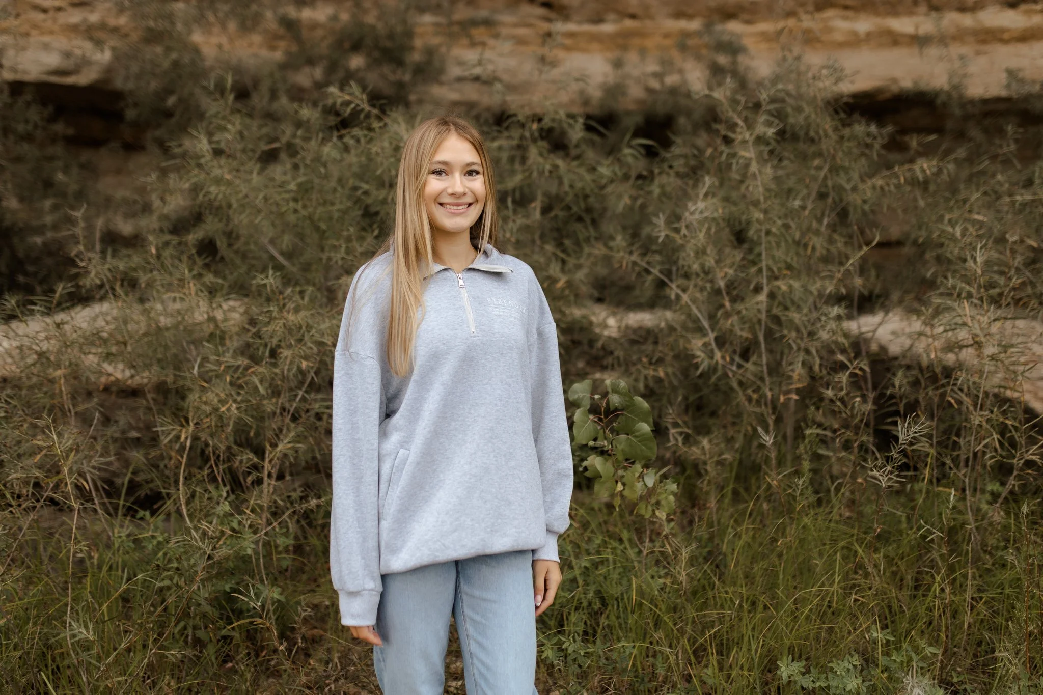 A young woman with long blonde hair, wearing a light gray sweatshirt and blue jeans, standing outdoors in front of a backdrop of tall grass and bushes, smiling at the camera.