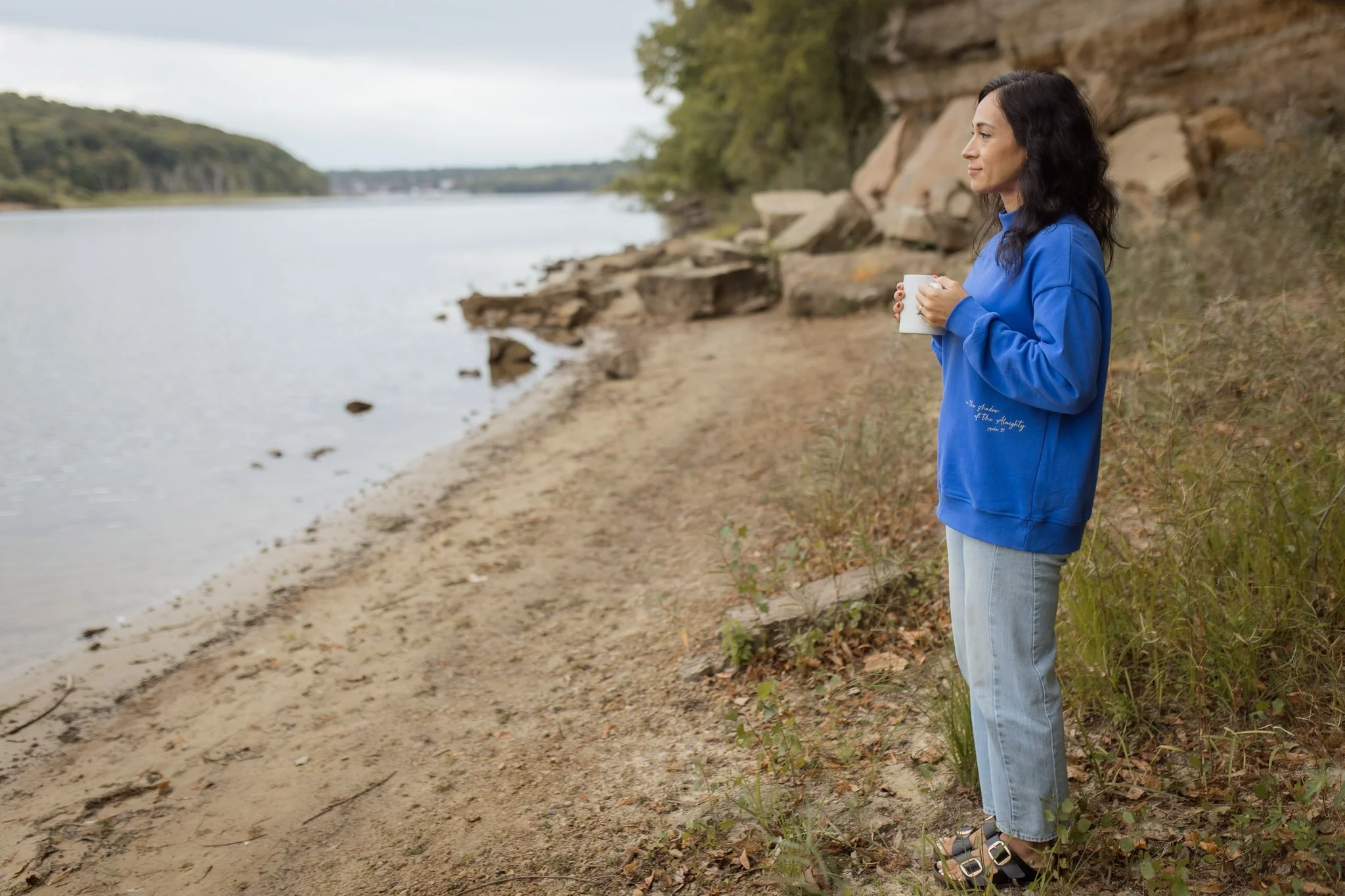 Woman standing by a river holding a white mug, wearing a blue jacket and jeans, with a rocky and wooded shoreline in the background.