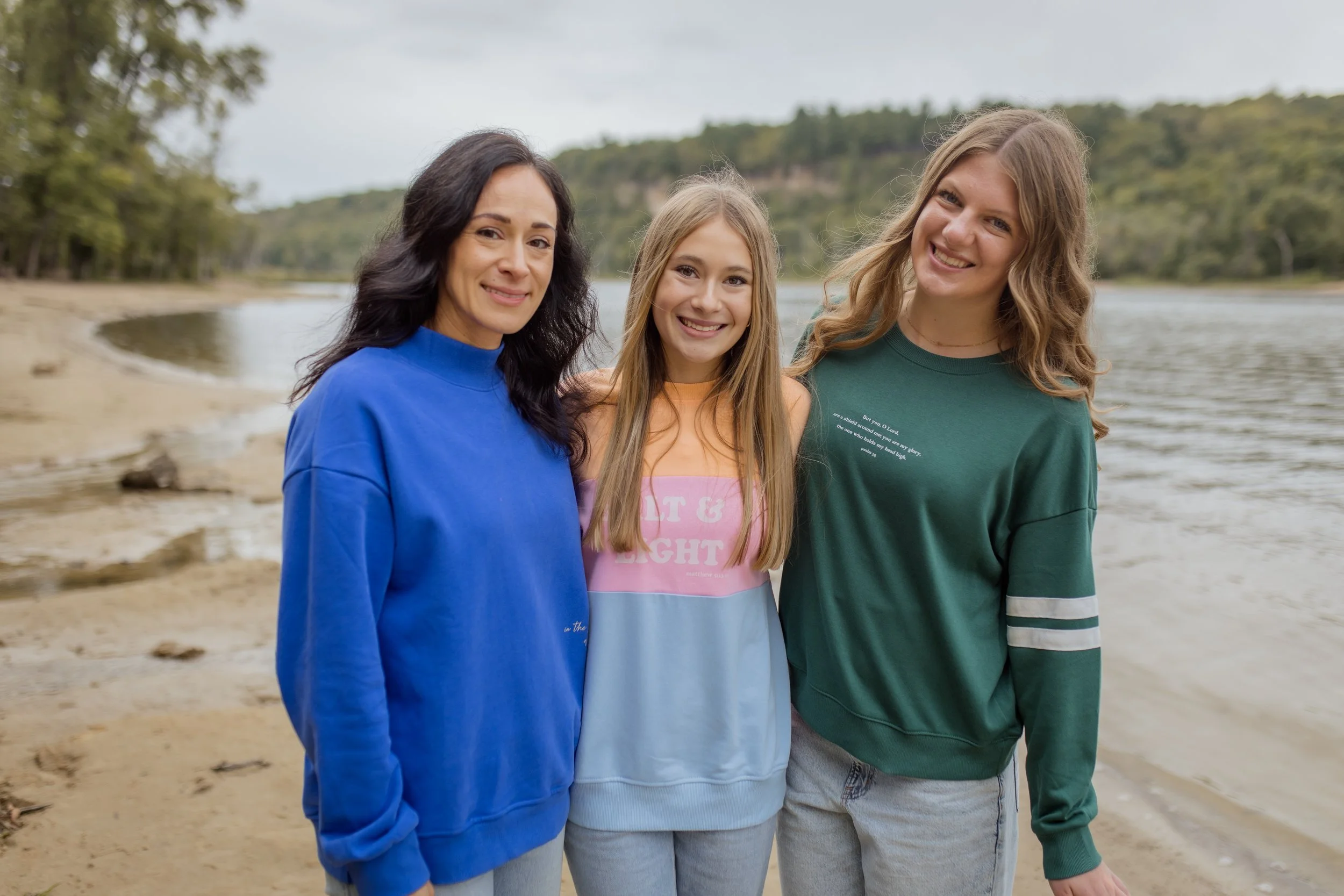 Three women, two adults and a girl, standing by a river with trees and a hill in the background, smiling at the camera during a christian fall line photoshoot.