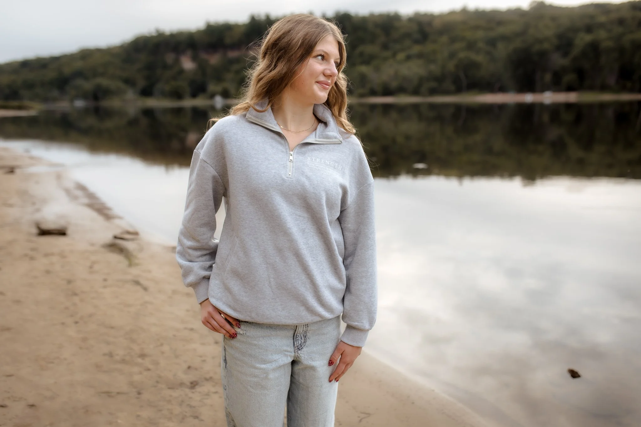 A young woman with wavy brown hair stands on a sandy beach near a calm lake, wearing a light gray quarter-zip sweatshirt and light jeans, with her hand resting on her hip, looking to the side with a serene expression.