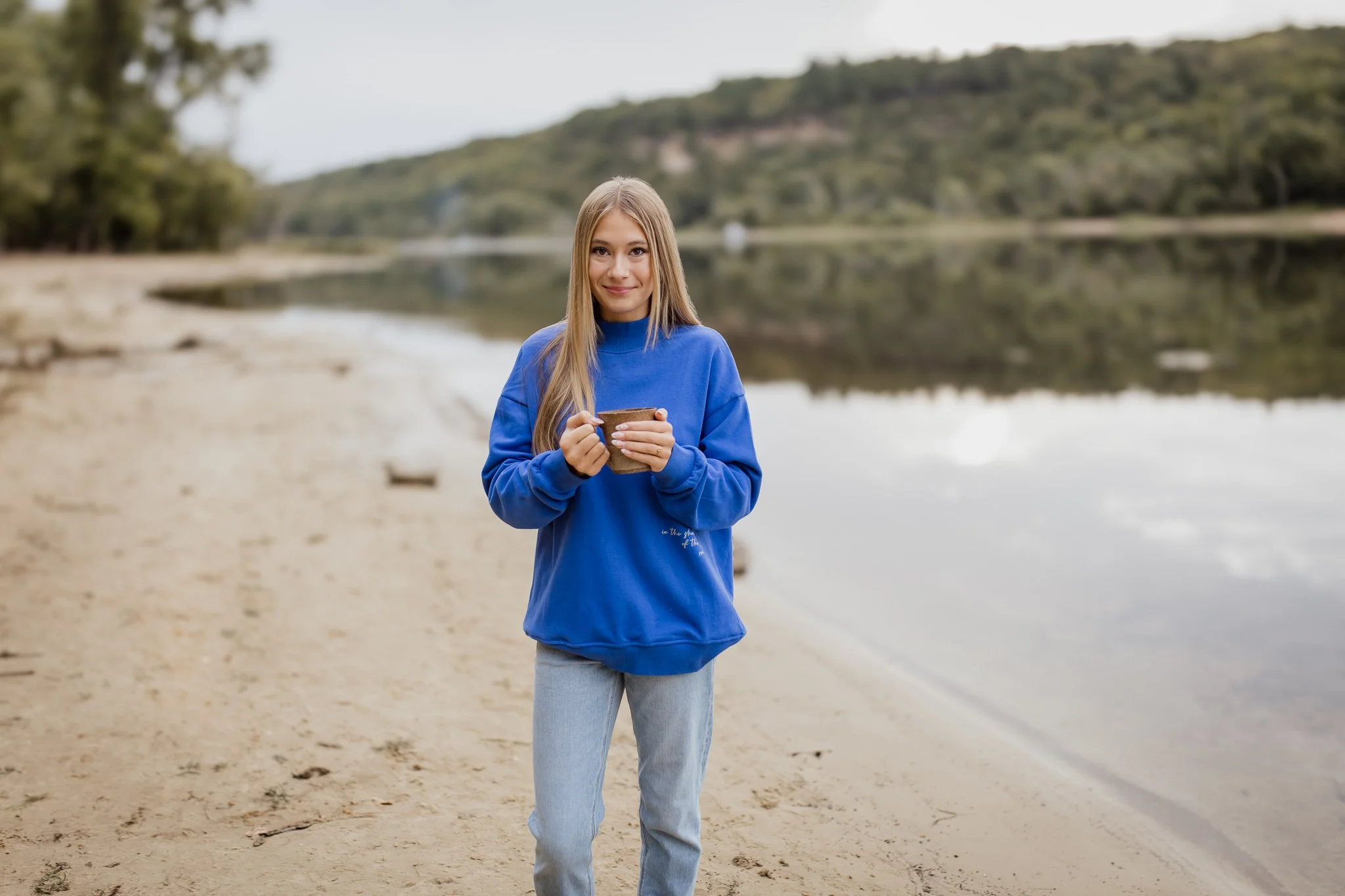 A young woman with long blonde hair is standing on a sandy beach near a body of water, holding a mug and smiling. She is dressed in a blue hoodie designed with bible verse psalm 91 shadow of the Almighty and light-colored jeans.