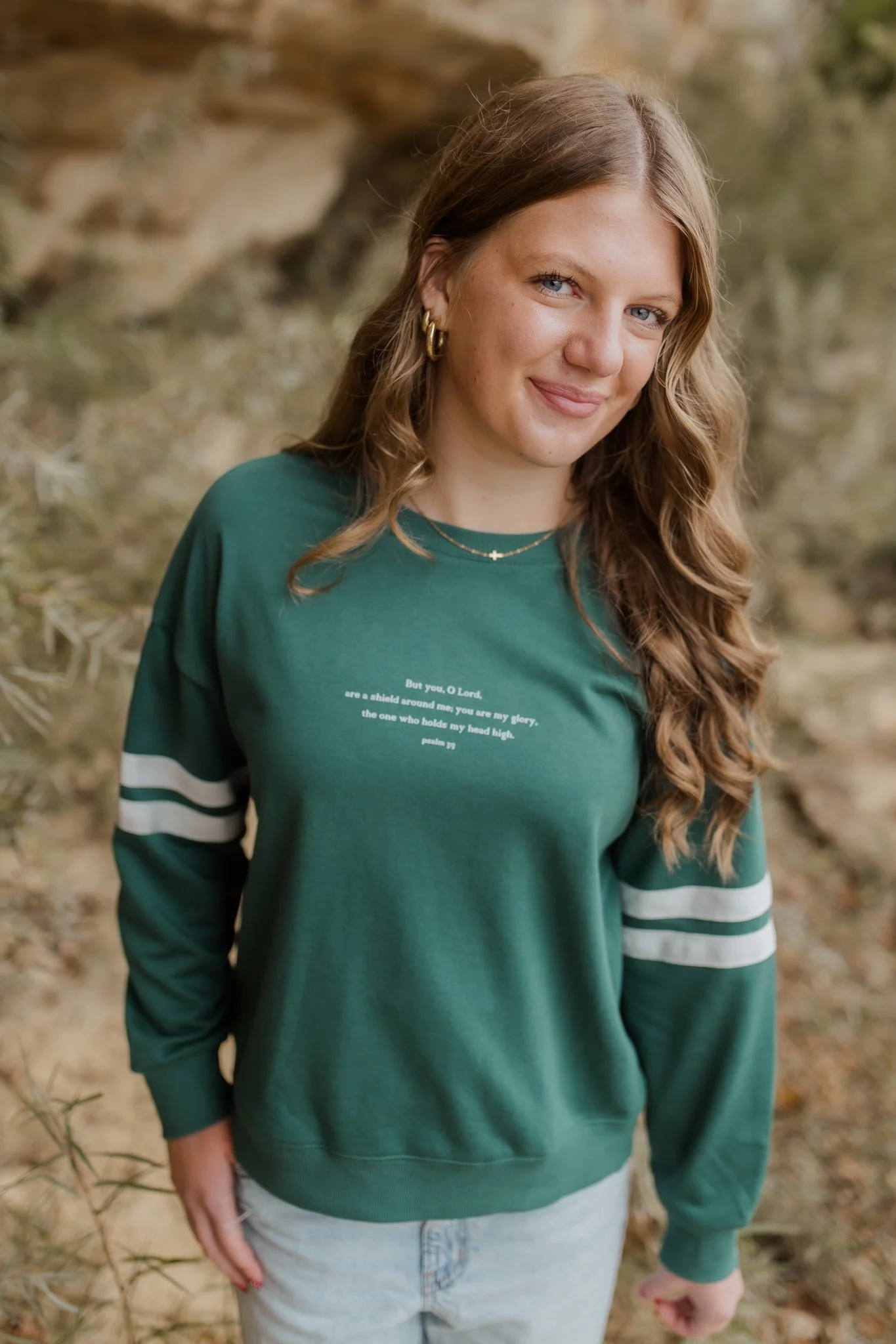 Woman wearing a green sweatshirt with white stripes on sleeves, gold hoop earrings, layered necklaces, and light-colored jeans, outdoors with a blurred natural background.