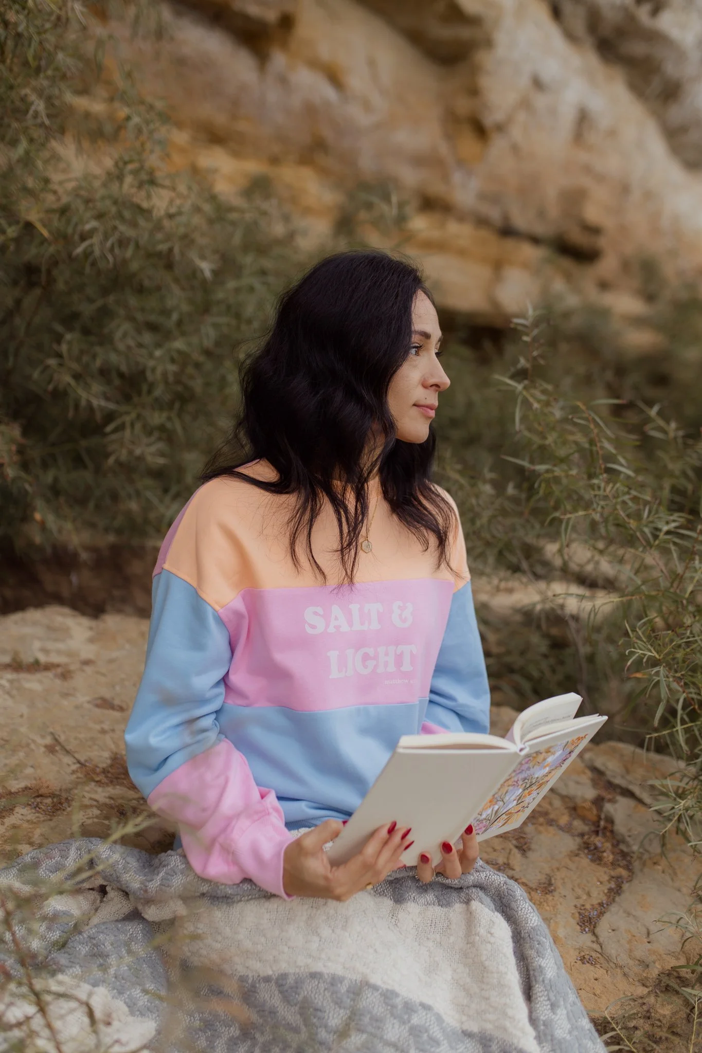 Woman with dark hair sitting outdoors on a blanket, reading a book, surrounded by plants and rocky terrain featuring a colorblock sweater with a bible verse Salt and Light to remind us that we are the salt of the earth and the light of the world.