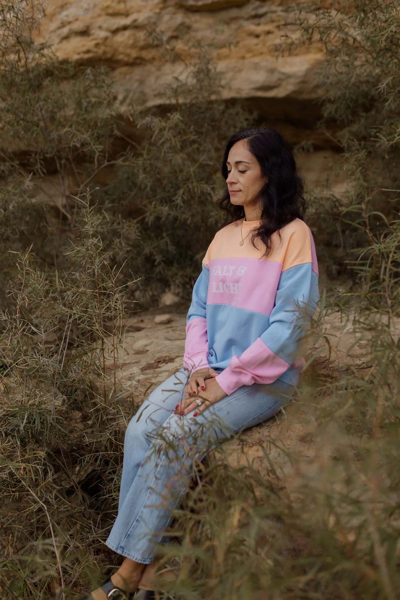 A woman kneeling on the ground in a natural outdoor setting with dry bushes and rocks, wearing a colorful pastel sweatshirt and light blue jeans, with dark wavy hair and a serene expression.