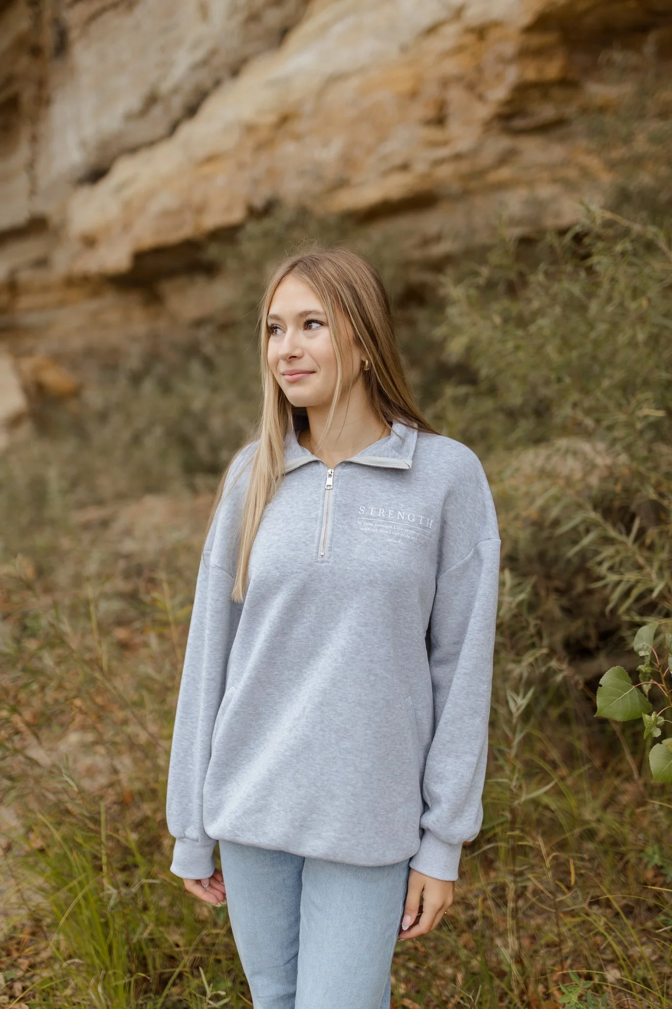 A young woman with long blonde hair standing outdoors in front of a rocky hillside and some greenery, wearing a grey zip-up sweatshirt and light blue jeans, looking to her left and smiling softly.
