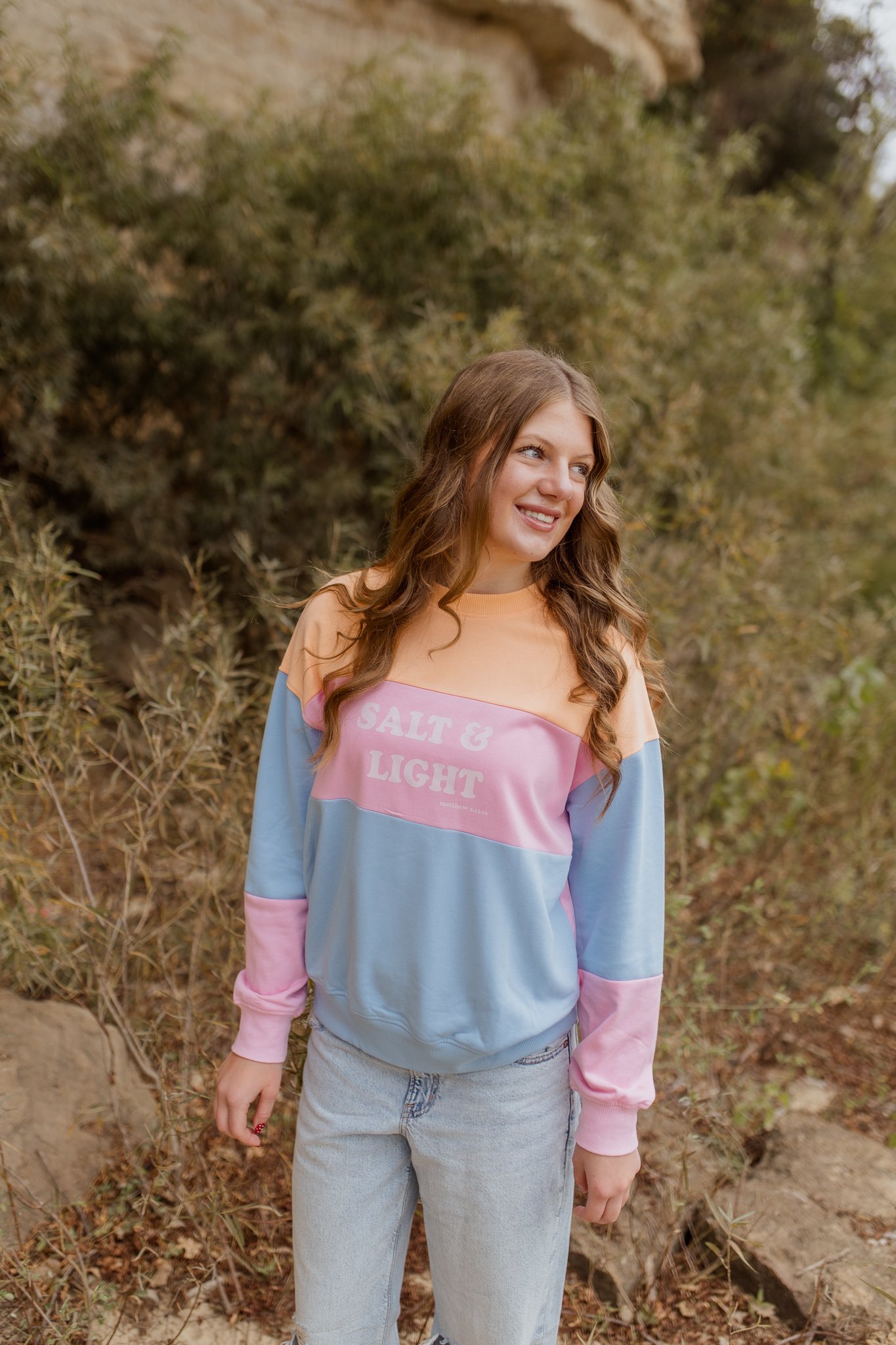 A young woman with long, wavy brown hair, wearing a pastel-colored sweatshirt with the words "Salt & Light" printed on it, standing outdoors in front of a background of trees and shrubs.