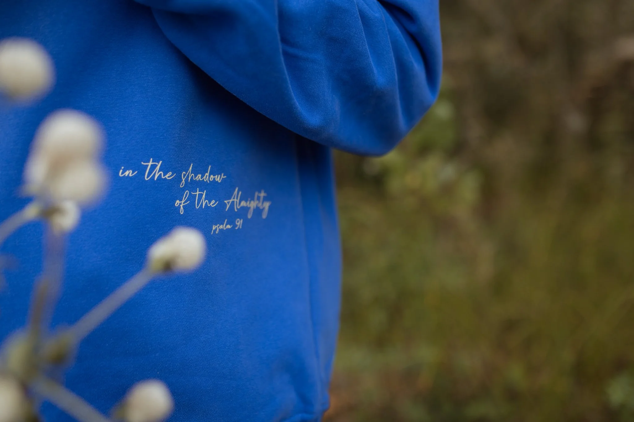 Close-up of a blue fabric with bible verse shadow of the almighty psalm 91, outdoors, with blurred white flowers in the foreground