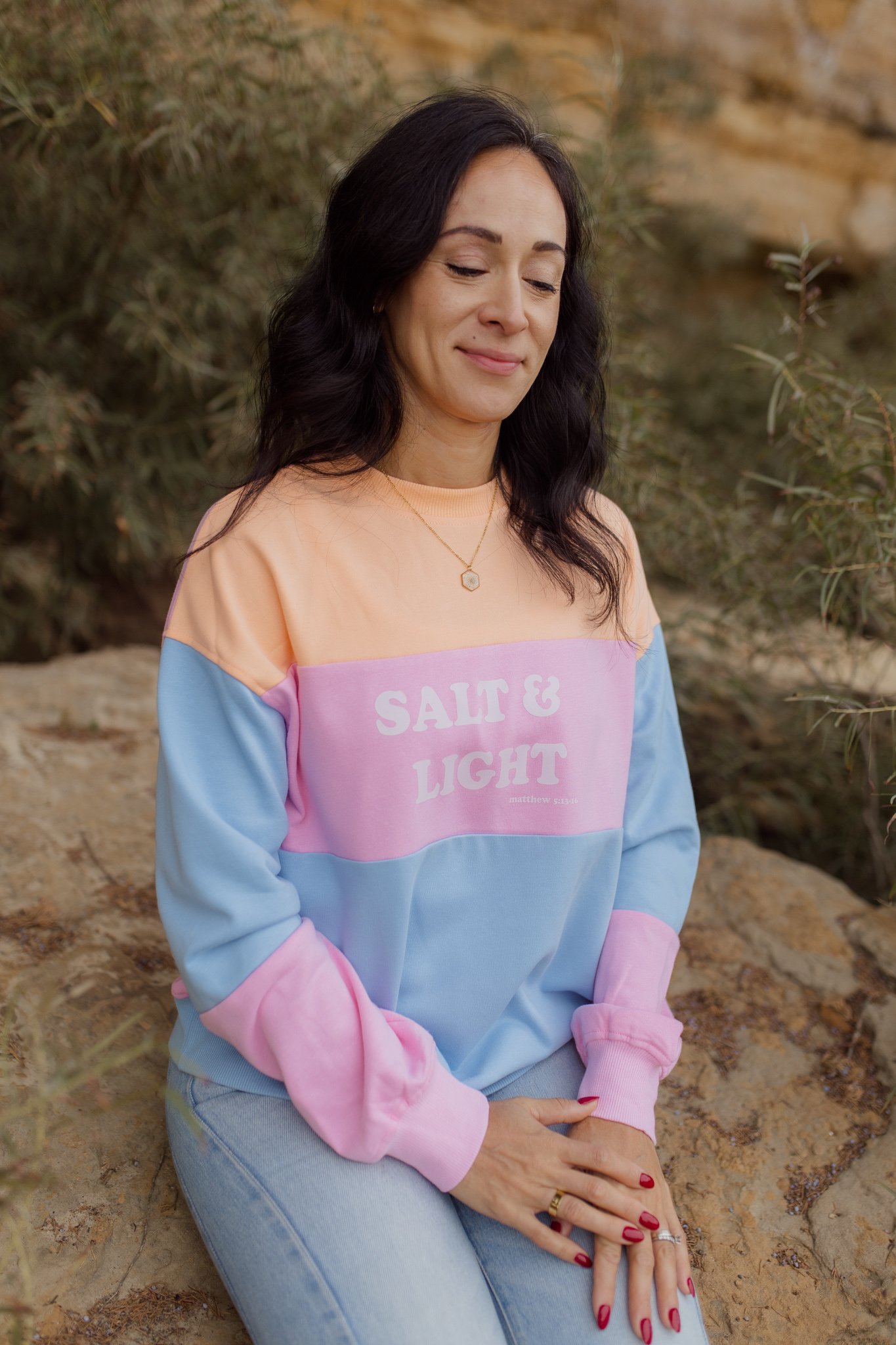 A woman with dark wavy hair and light skin, wearing a pastel-colored sweatshirt with pink, peach, and blue sections with design Salt and Light bible verse, and light blue jeans, is sitting outdoors on a rocky ground with greenery in the background. 