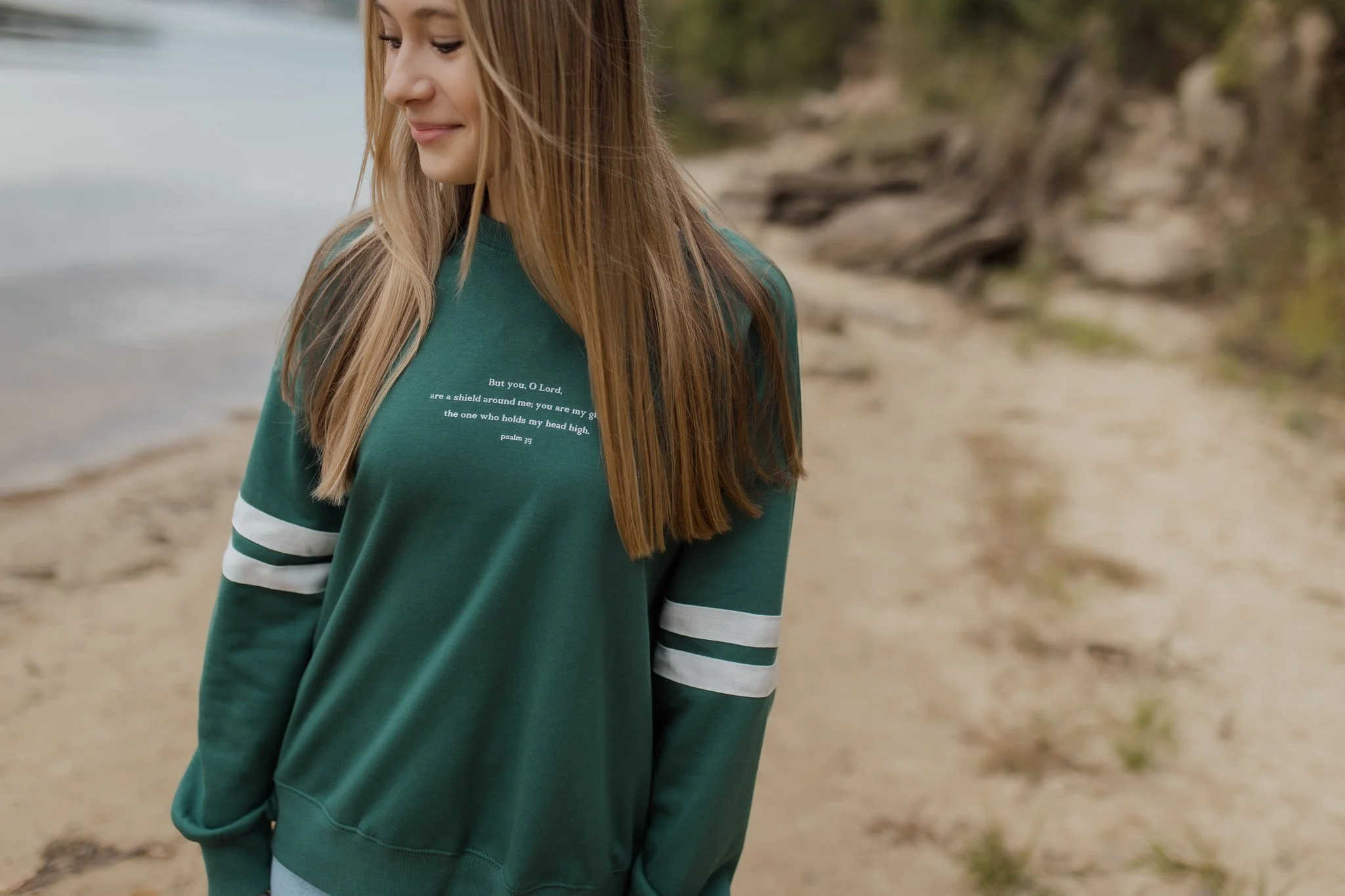 A young woman with long blonde hair stands near a boat on a sandy beach, wearing a green sweatshirt with white and black stripes on the sleeves. The sweatshirt has a Bible verse printed on it from Psalm 33.
