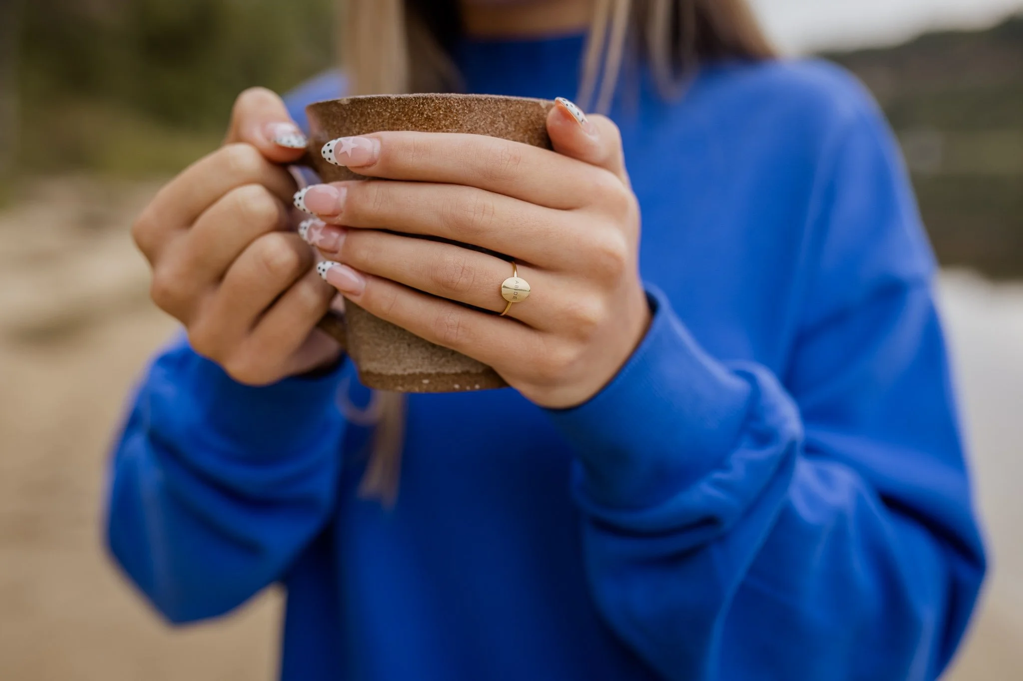 Person wearing a blue jacket holding a brown ceramic mug with both hands, nails painted with white, black, and pink stars and dots, wearing a gold ring with the word 'ADIDE' engraved on it.