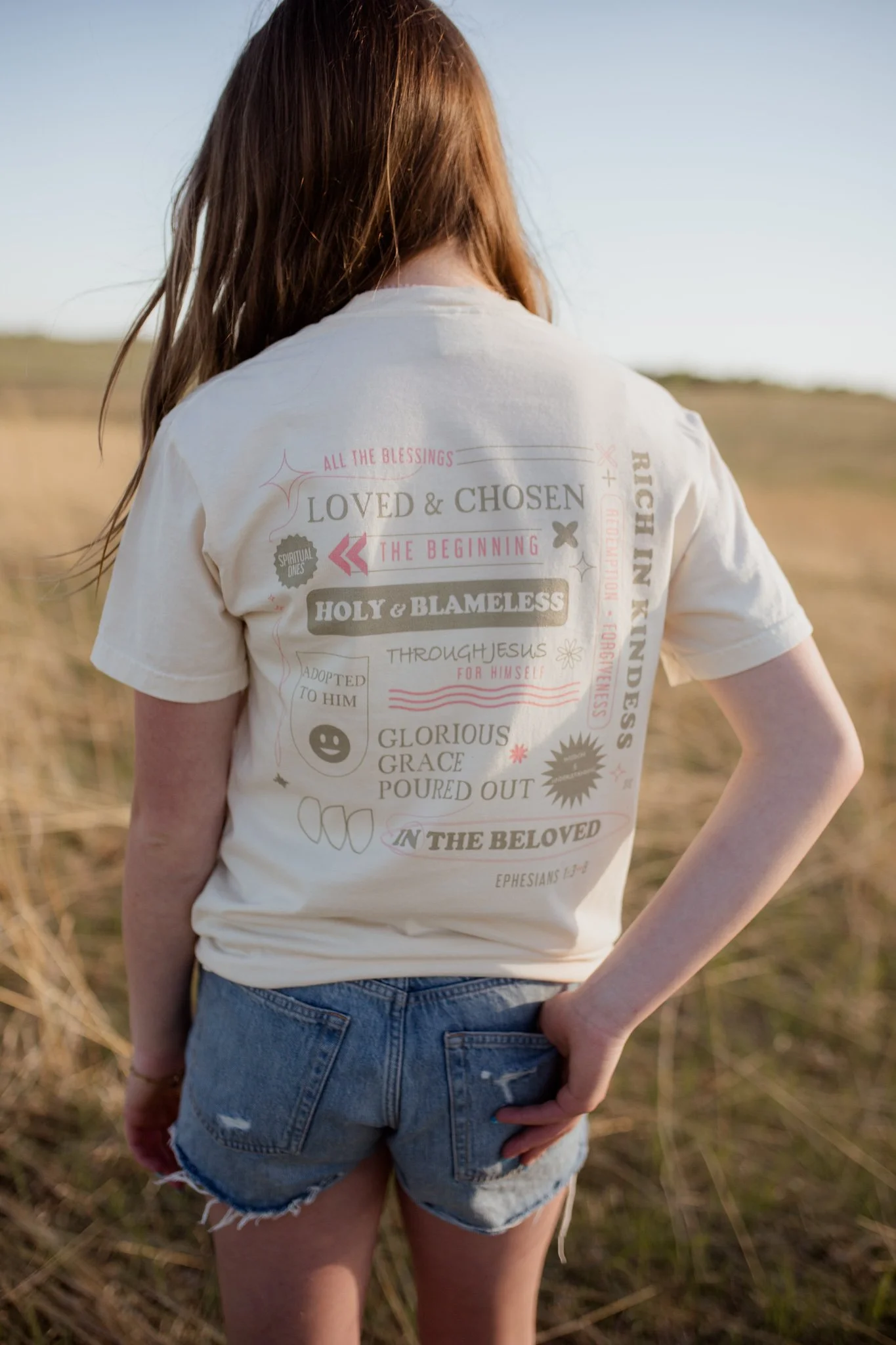 Christian comfort colors True Identity in Christ T-shirt inspired by bible verse Ephesians 1:3-8.  ivory tee with pink and tan screen print reminding believers that they are deeply chosen and loved.