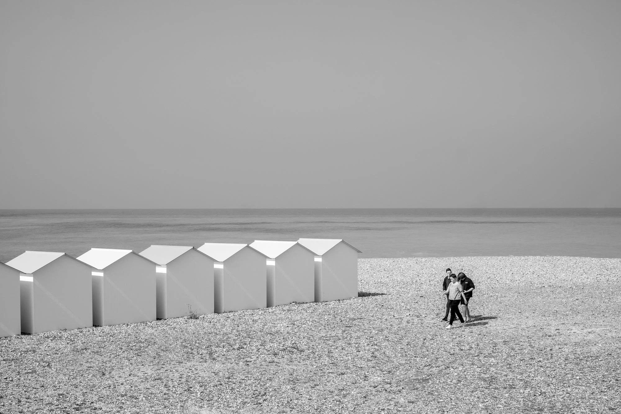  Black-and-white seaside photograph on the beach at Le Tréport, Normandy, three friends walking by the sea near a row of beach huts, by Chris Perona. 