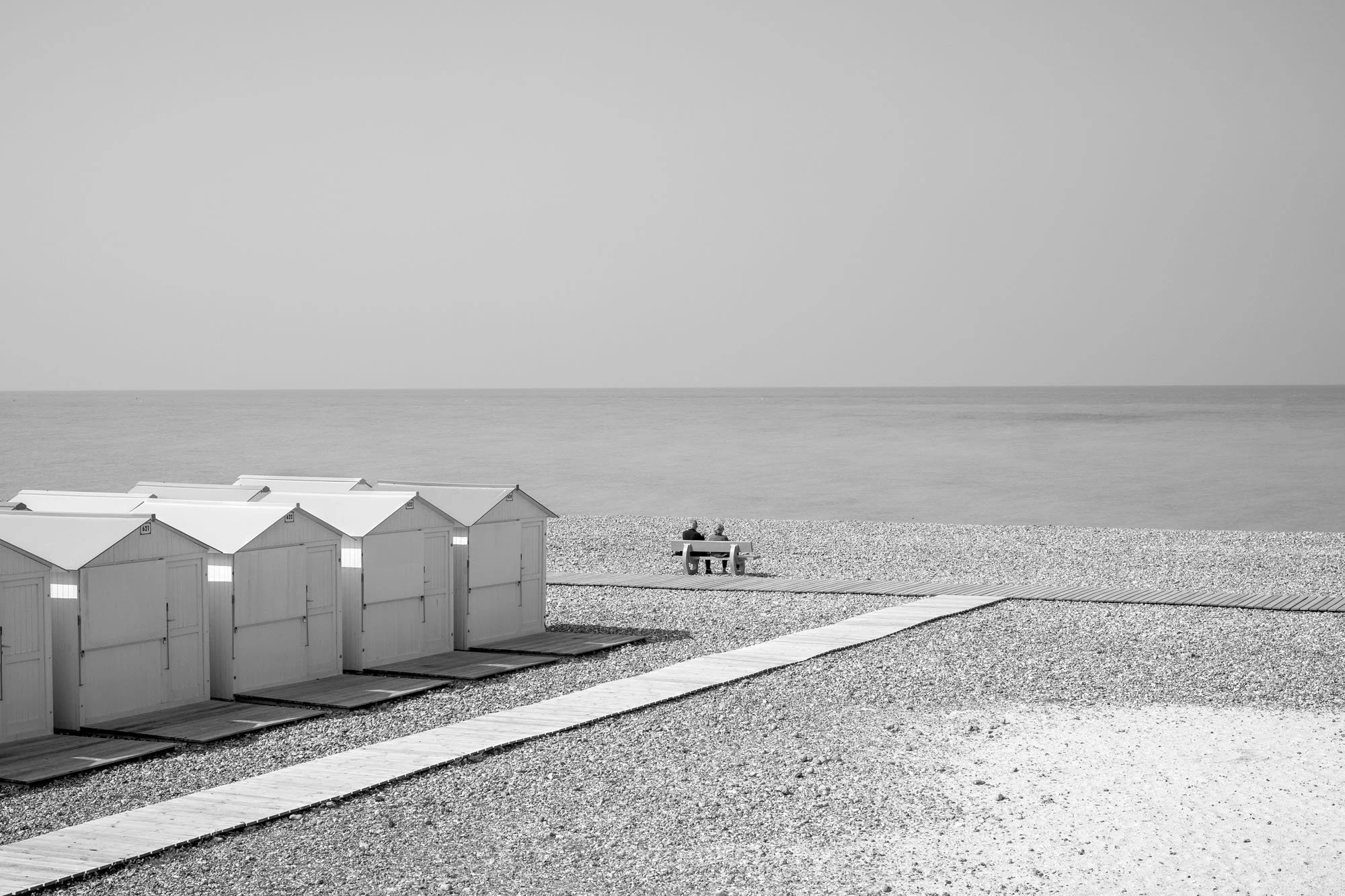  Black-and-white seaside photograph on the beach at Le Tréport, Normandy, beach huts and a couple seated facing the sea, by Chris Perona. 