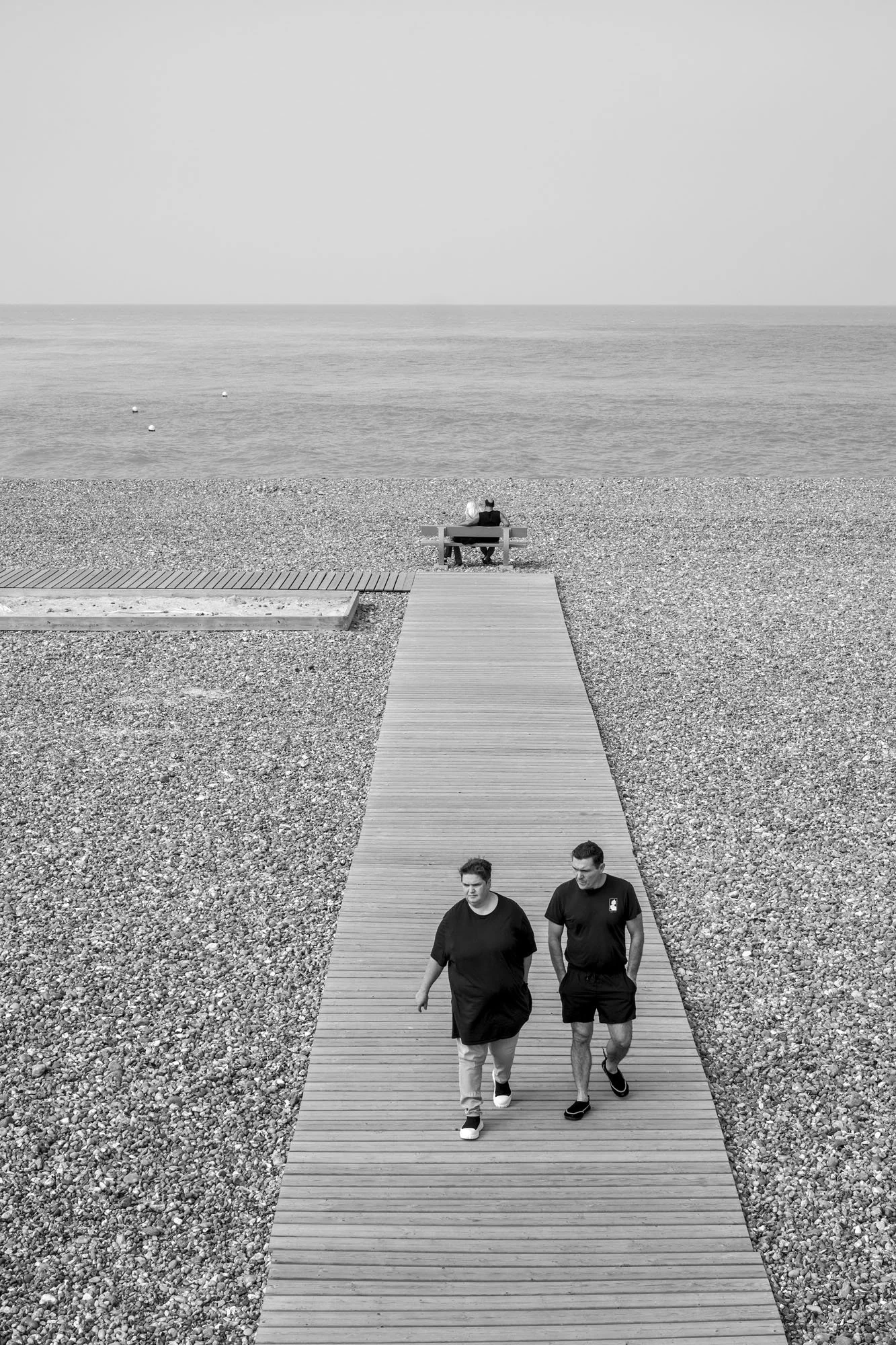  Black-and-white seaside photograph on the beach at Le Tréport, Normandy, a couple seated facing the sea, a father and son walk towards the camera, by Chris Perona. 