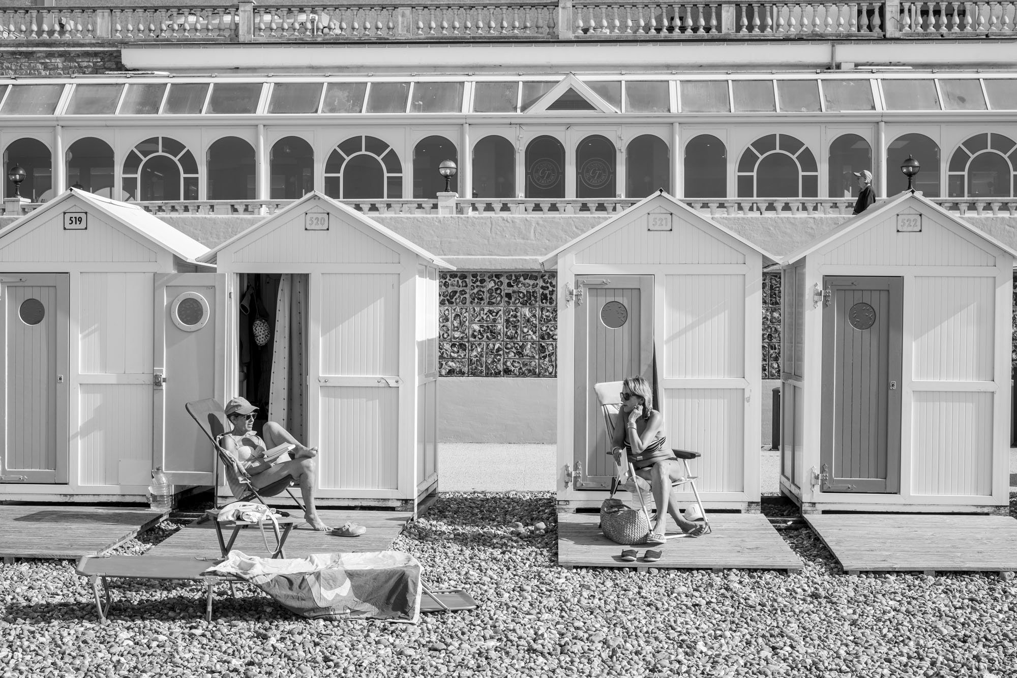  Black-and-white seaside photograph on the beach at Le Tréport, Normandy, two women chatting while sunbathing by their beach huts by Chris Perona. 