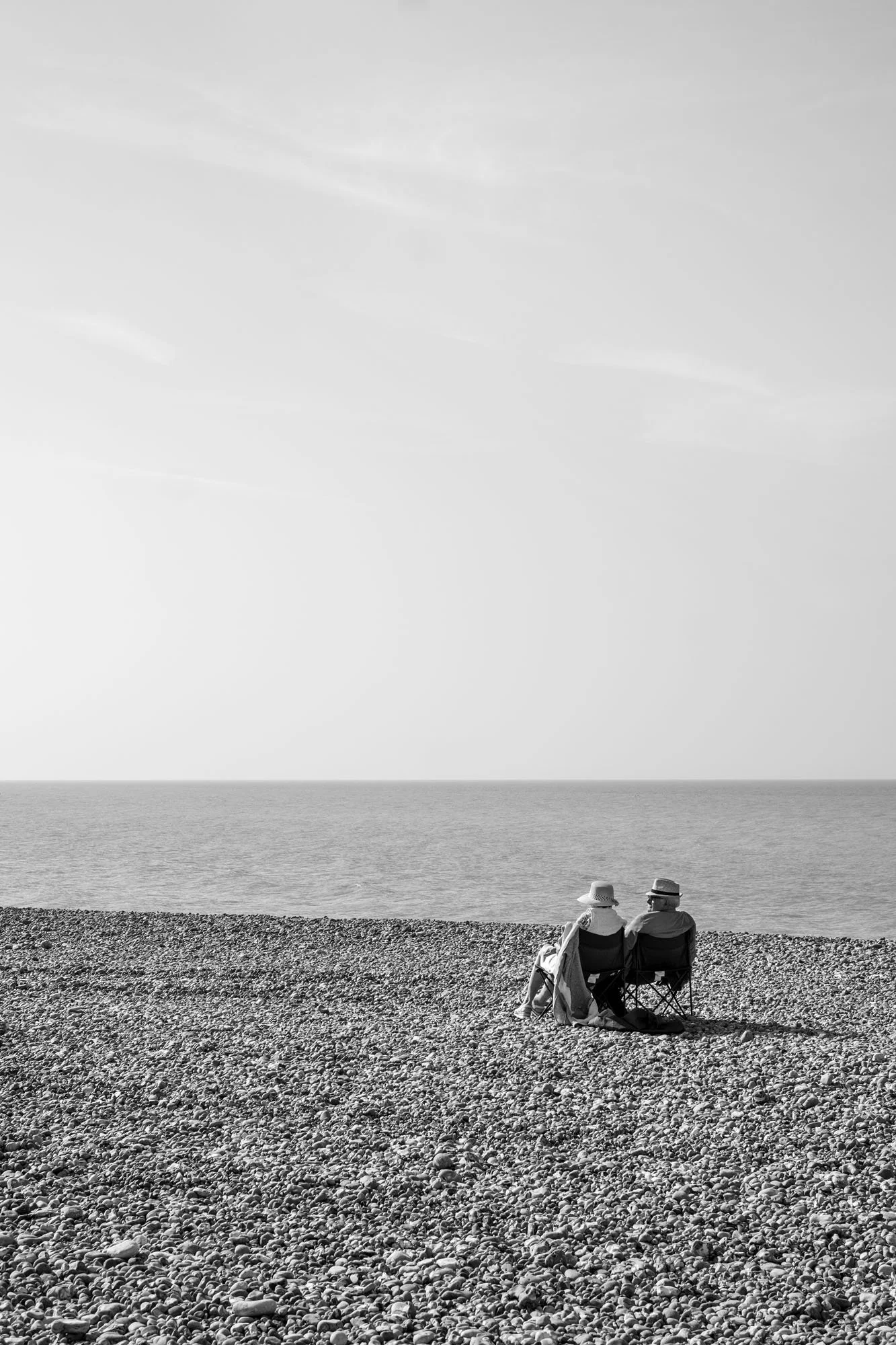  Black-and-white seaside photograph on the beach at Le Tréport, Normandy, a lonely senior couple sunbathing facing the sea on a pebbles beach, by Chris Perona. 