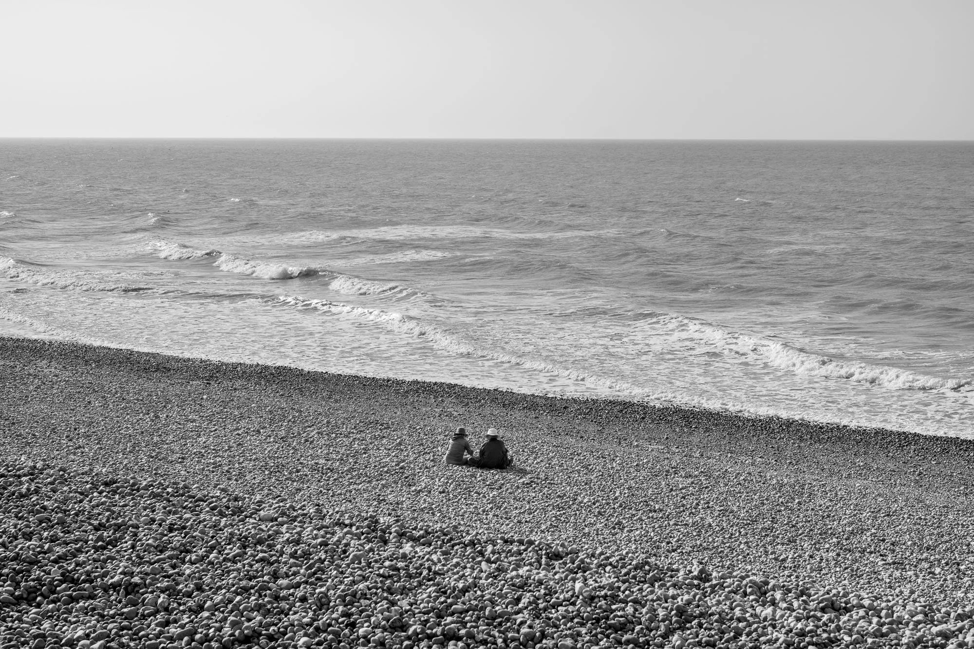  Black-and-white seaside photograph on the beach at Le Tréport, Normandy, two people seated at a pebble beach facing the sea, by Chris Perona. 